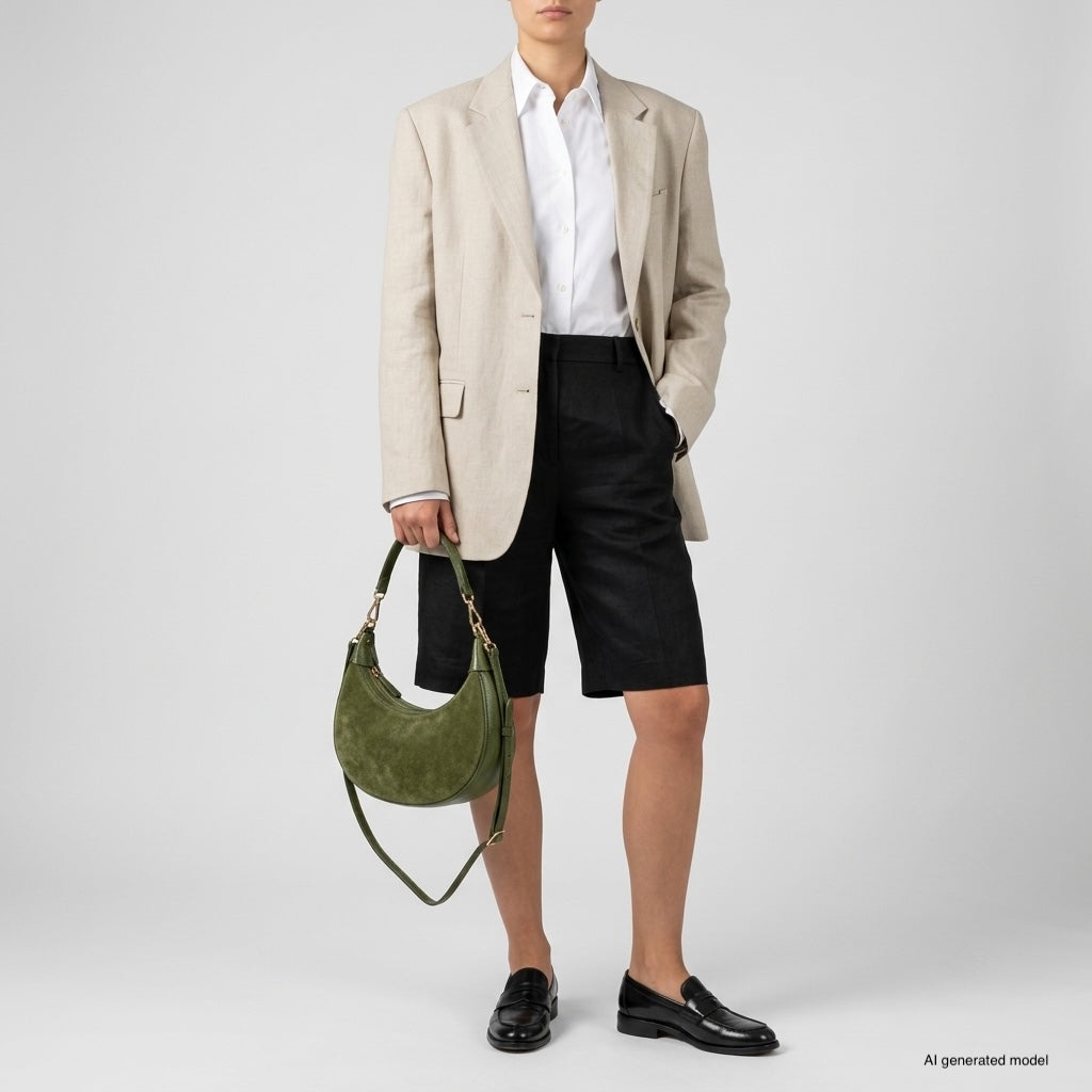 A person wearing a beige blazer, white shirt, black knee-length shorts, and black loafers holds an elegant Italian leather bag against a plain background.
