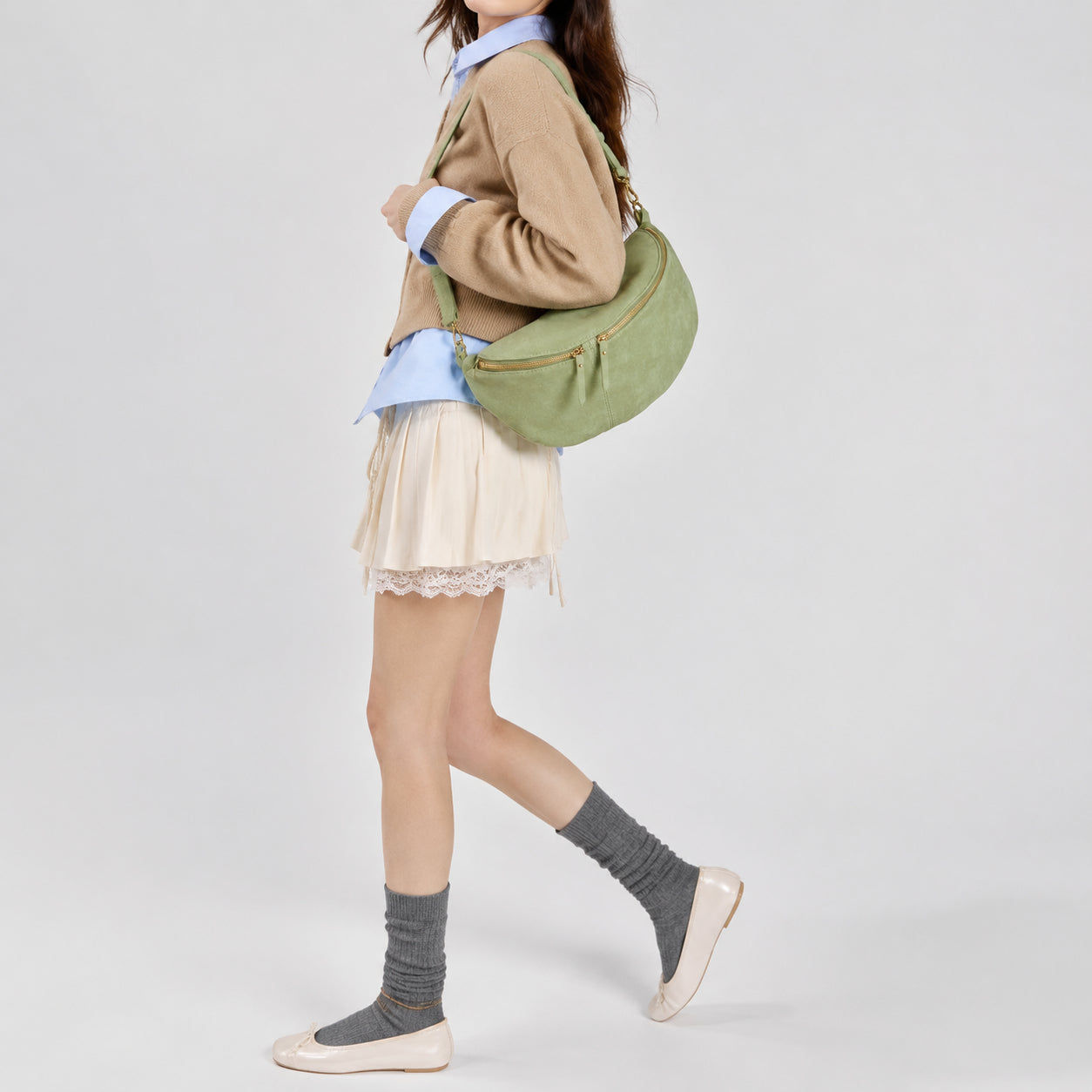 A woman walks wearing a tan sweater over a blue shirt, a cream pleated skirt with lace trim, gray socks, beige flats, and an elegant Florentine leather bag against a plain background.