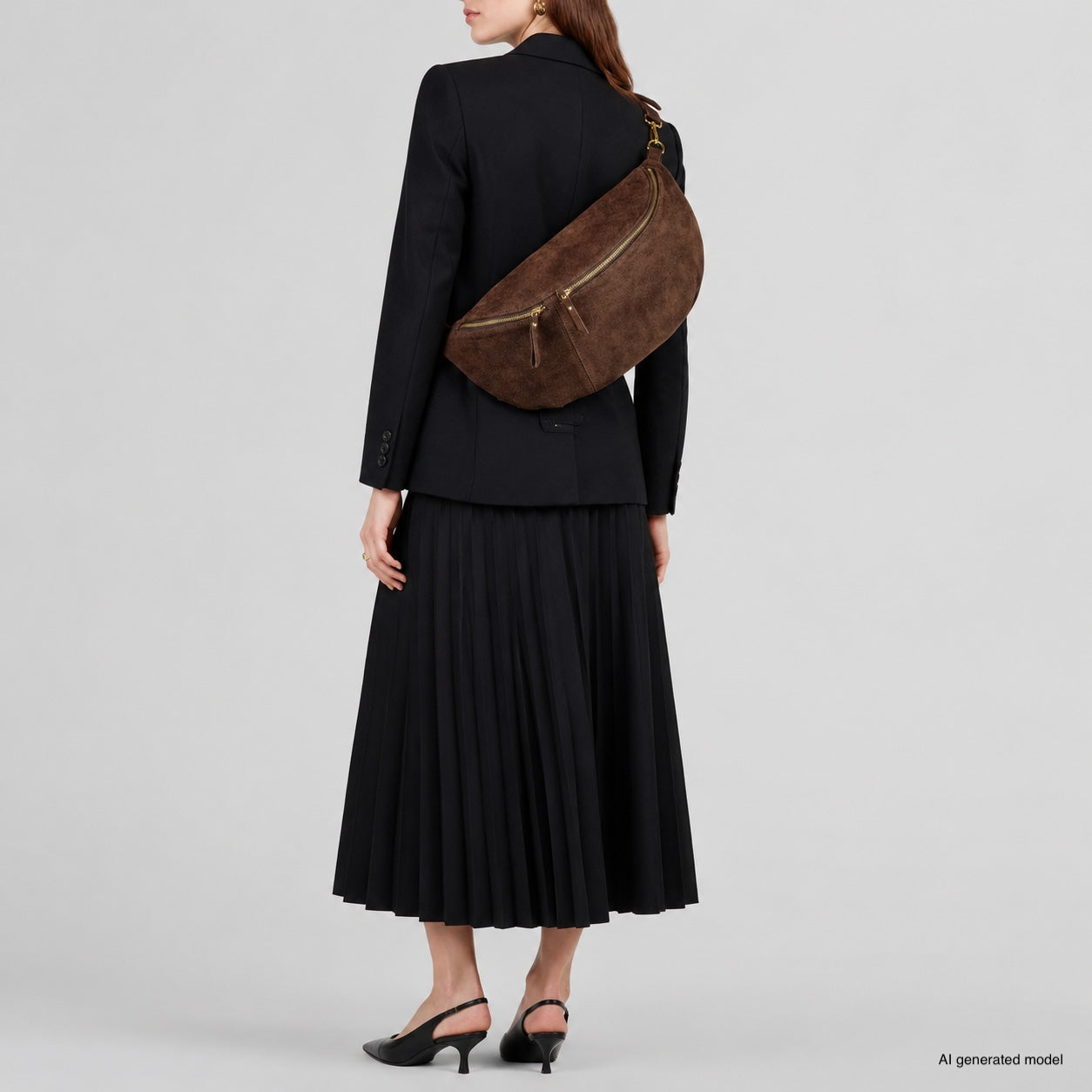 A woman wearing a black blazer, pleated skirt, and black heels stands facing away, carrying a large Italian suede crossbody bag over her shoulder. The background is plain and light-colored.