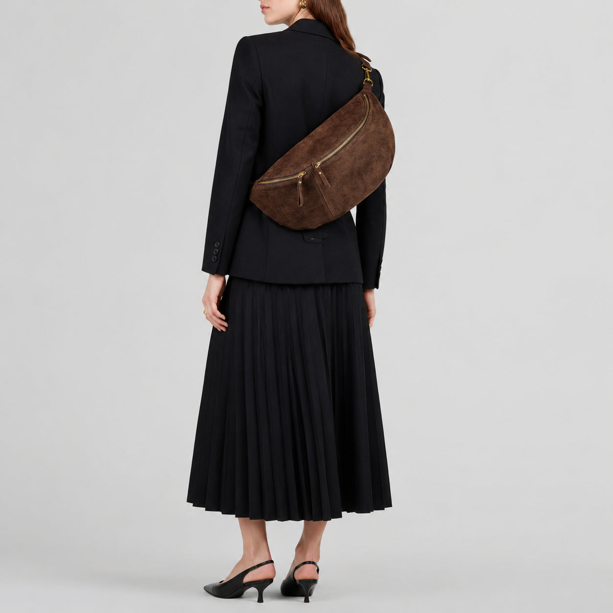 A woman in a black blazer and pleated skirt stands facing away, wearing black heels and carrying an Italian suede crossbody over her shoulder.