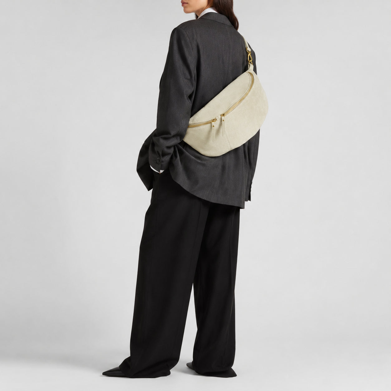 A person in an oversized black suit with wide-leg pants and an oversized blazer, shown from behind, carries a large Florentine leather bag with gold zippers. The background is plain and light gray.