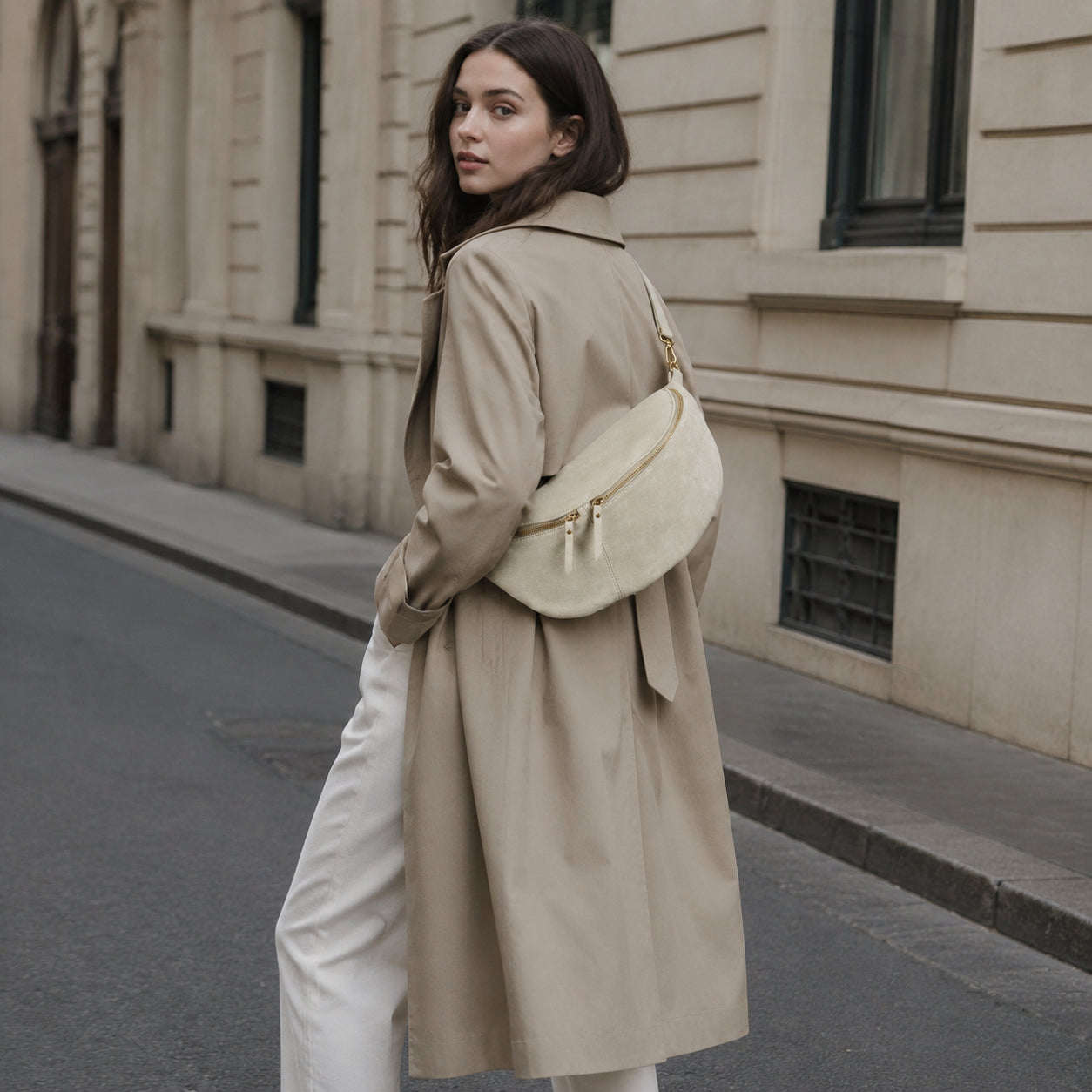 A woman with long brown hair wearing a beige trench coat, white pants, and an Italian suede crossbody stands on a city street, looking over her shoulder. Stone buildings line the sidewalk in the background.