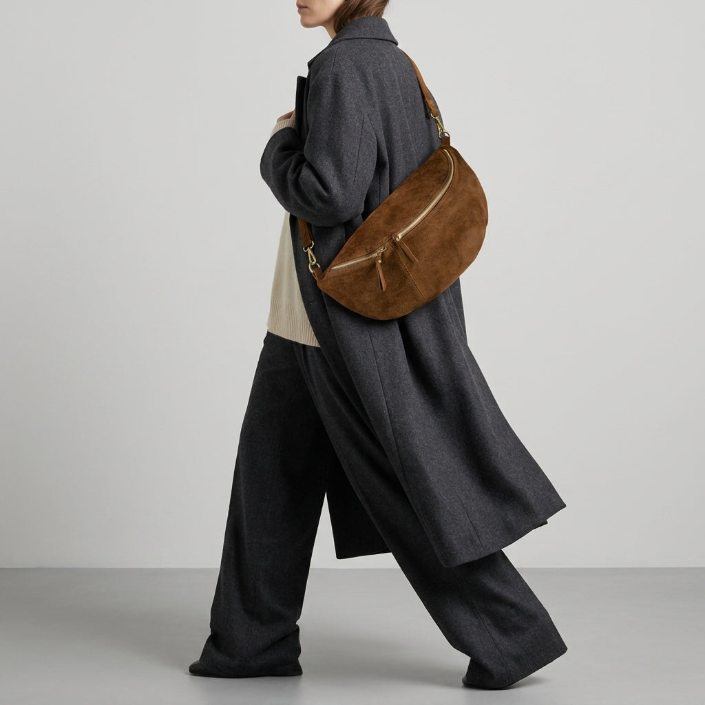 A person wearing a long dark grey coat and wide-leg pants walks with an Italian suede crossbody bag over their shoulder against a plain light background.
