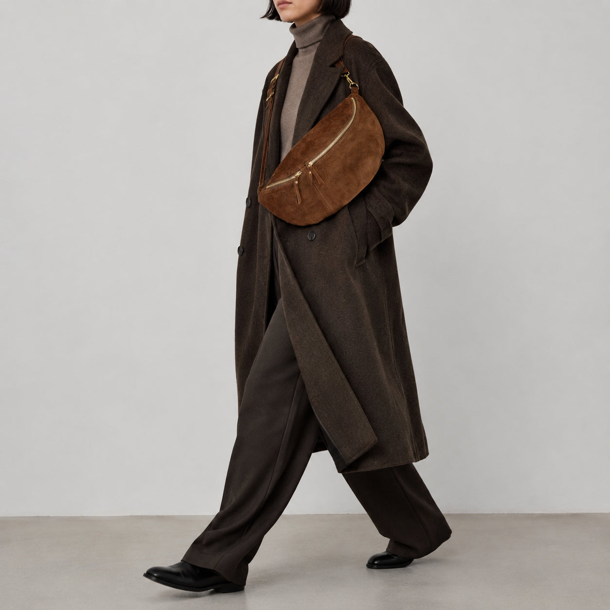 A person wearing a long brown coat, brown turtleneck, wide-leg trousers, and black shoes carries an Italian suede crossbody with gold hardware. The background is plain light gray.