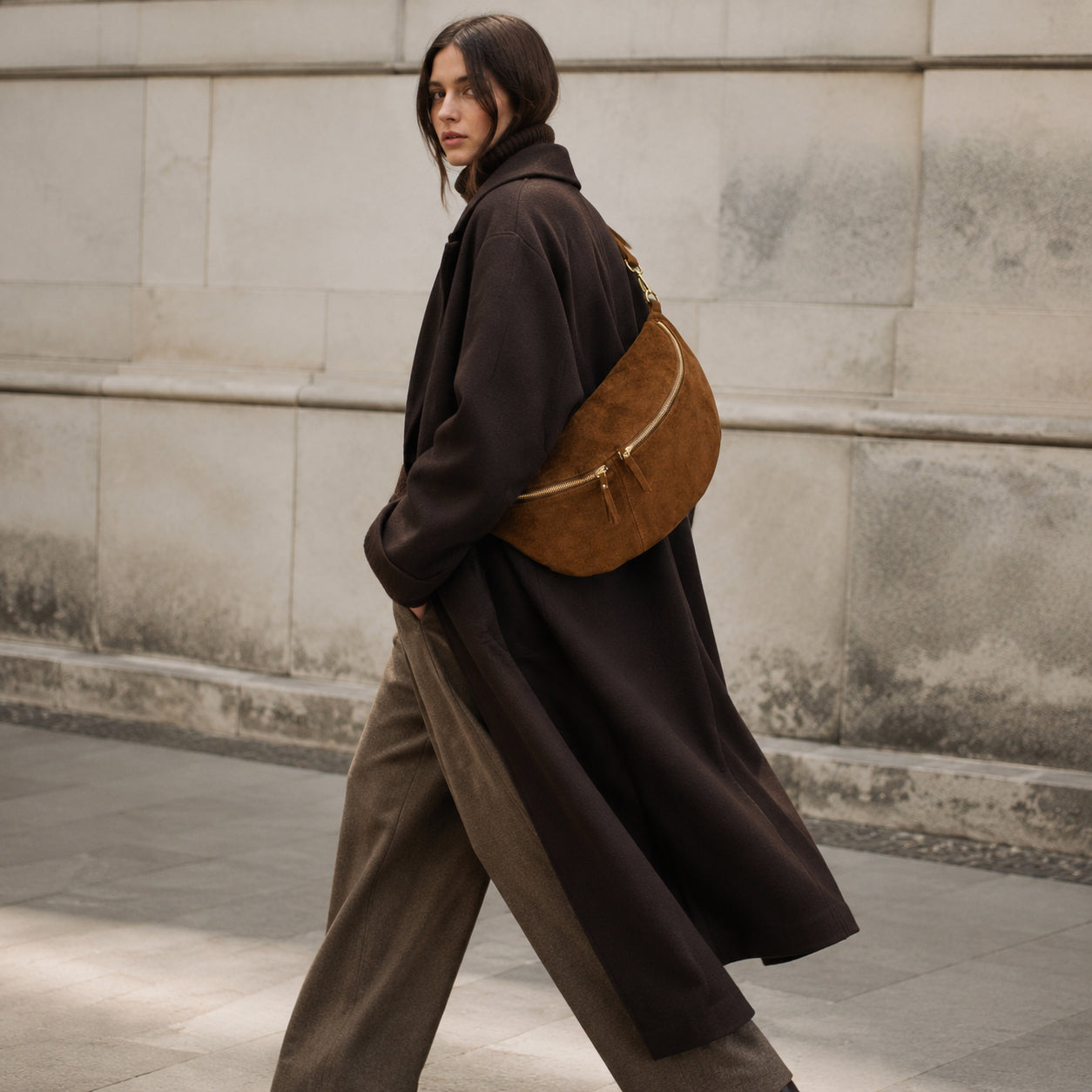 A person with long brown hair wears a long dark coat, brown wide-leg trousers, and carries an Italian suede crossbody bag while walking on a city sidewalk beside a light stone wall.