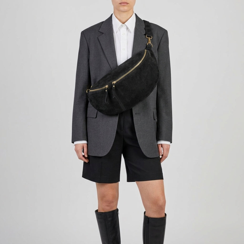 A person wearing a grey blazer, white shirt, black knee-length shorts, knee-high black boots, and a large Florentine leather bag stands against a plain light background.