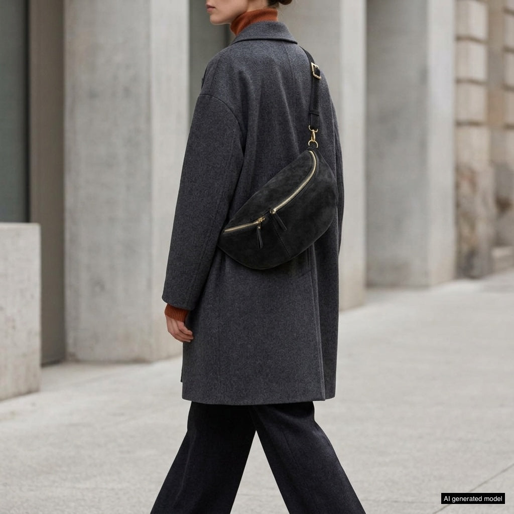 A person wearing a long dark gray coat and black pants carries an Italian suede crossbody bag while walking outdoors on a city sidewalk, their head turned slightly away from the camera.