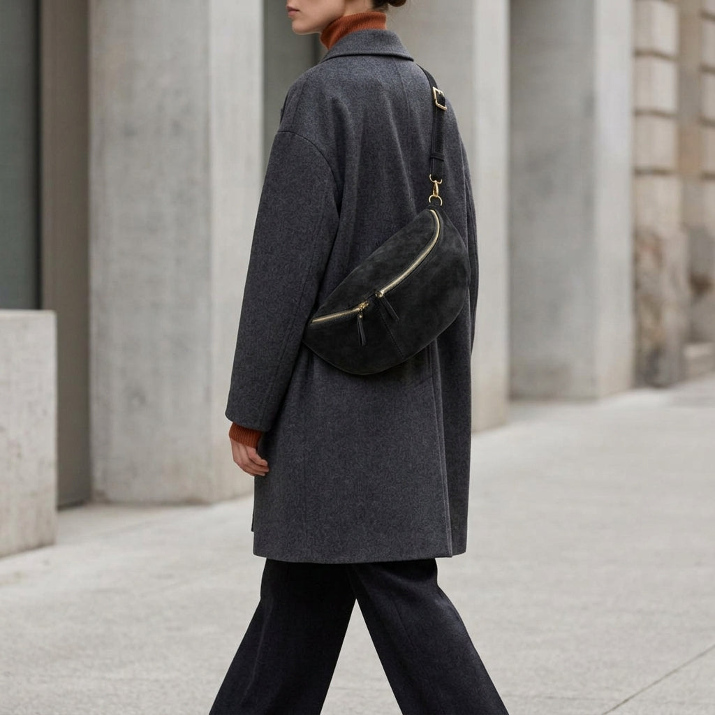 A person wearing a long dark gray coat, matching pants, and an Italian suede crossbody is walking outdoors along a modern, light-colored stone building. Their face is mostly out of frame.