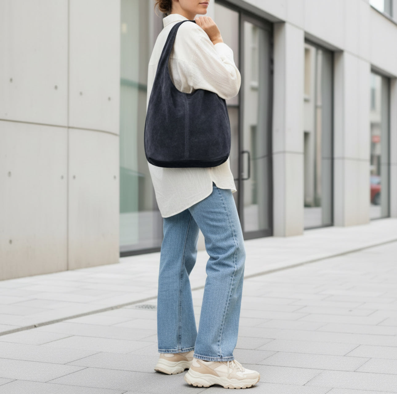 A person stands on a city sidewalk wearing light blue jeans, an oversized white shirt, chunky beige sneakers, and carrying a minimalist suede handbag. The background features modern buildings with large windows.