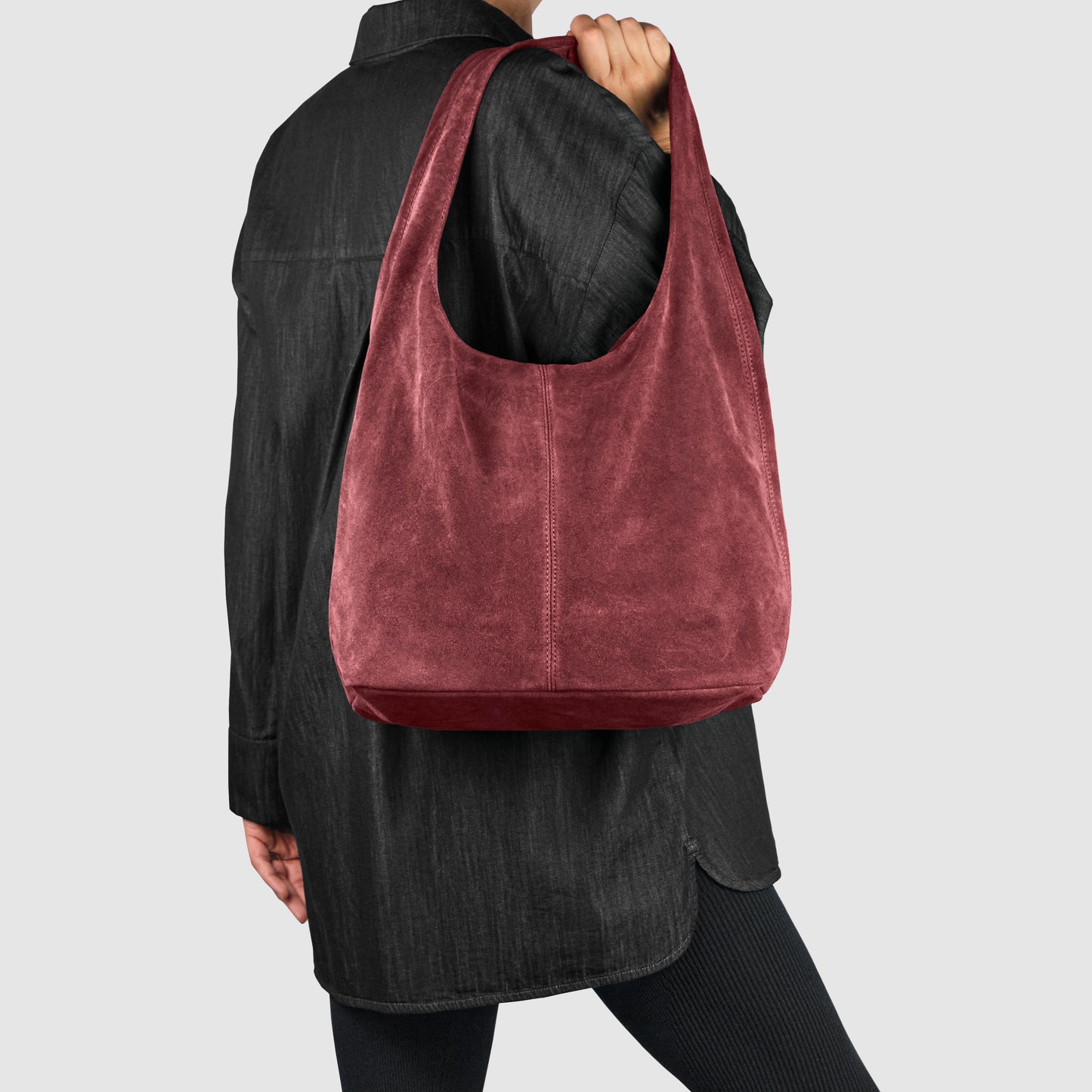 A person wearing a black oversized shirt and black leggings holds a large, soft Italian suede bag, showcasing refined Florentine craftsmanship, against a plain light gray background.