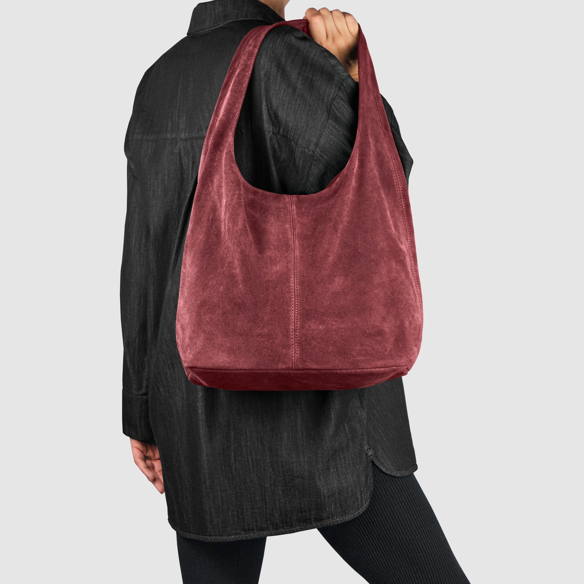 A person wearing a black oversized shirt and black leggings holds a large, soft Italian suede bag, showcasing refined Florentine craftsmanship, against a plain light gray background.