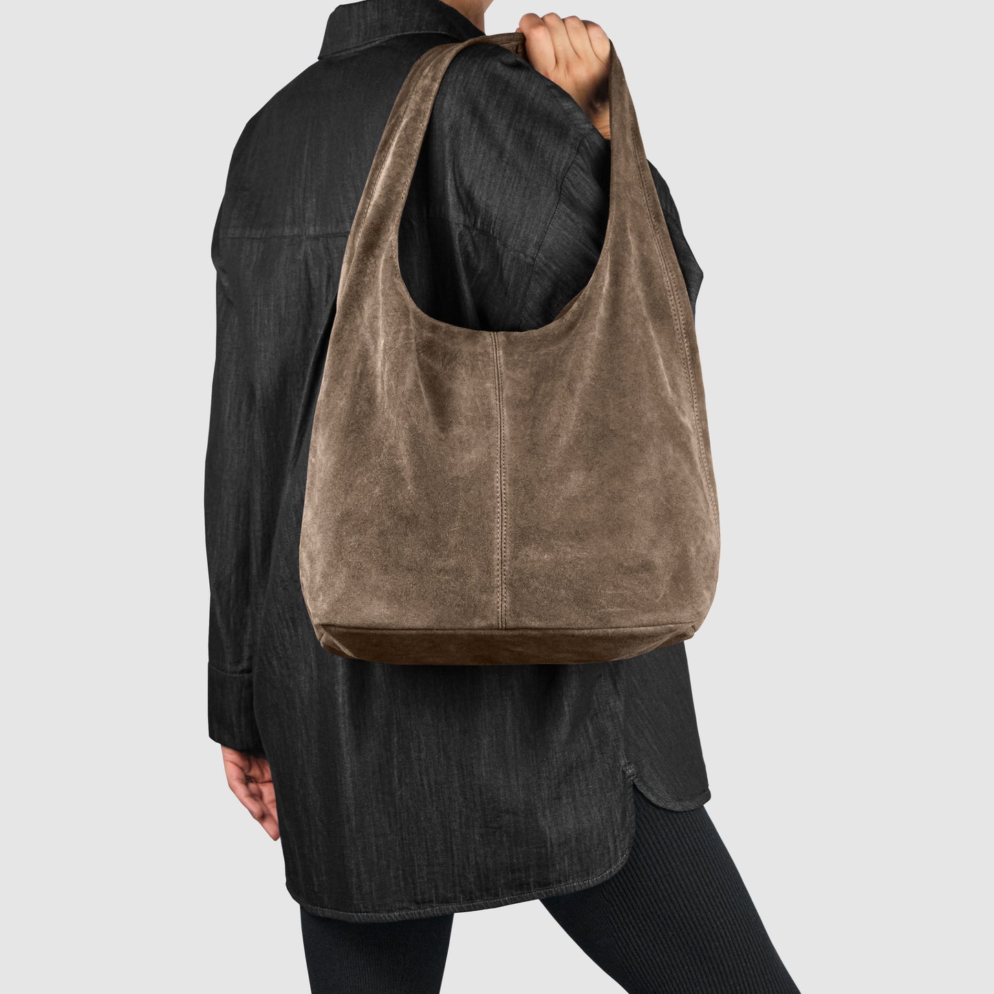 A person wearing a black jacket and pants holds a large, minimalist shoulder bag crafted from brown suede on their arm against a plain light background.