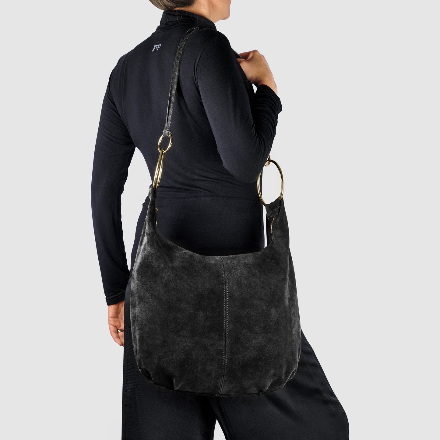 A person in a black outfit stands against a gray background, holding a large Italian handmade bag crafted from black natural suede with gold hardware over their left shoulder. The bag has a soft, slouchy shape.