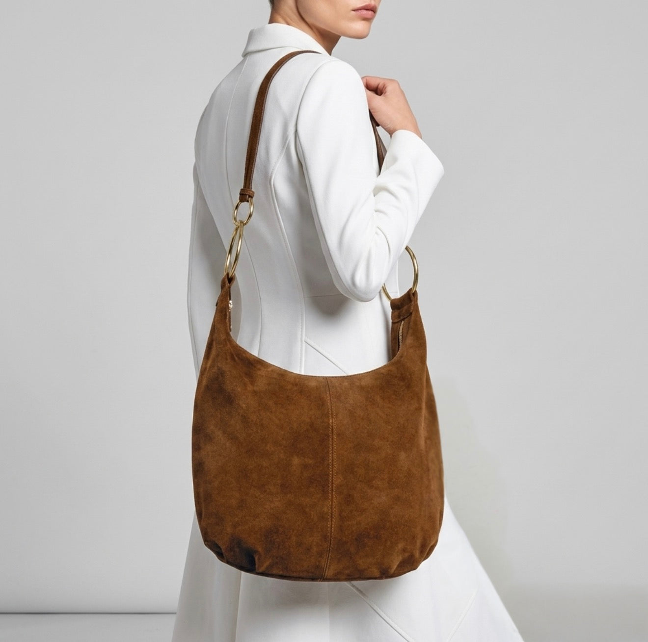A woman in a white coat is shown from the side, carrying a slouchy medium purse crafted from brown suede with gold hardware over her shoulder against a plain gray background. Her face is partially out of frame.