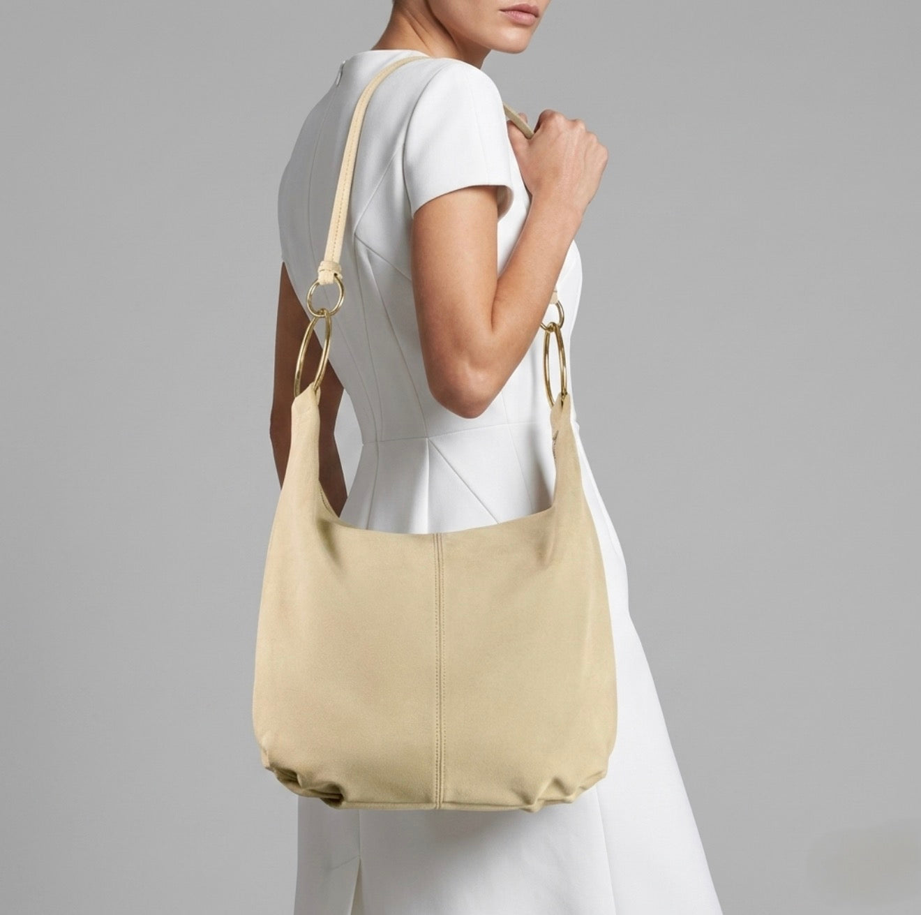 A woman in a white short-sleeve dress holds a handmade Italian bag with gold rings and a matching strap, posing against a plain gray background.