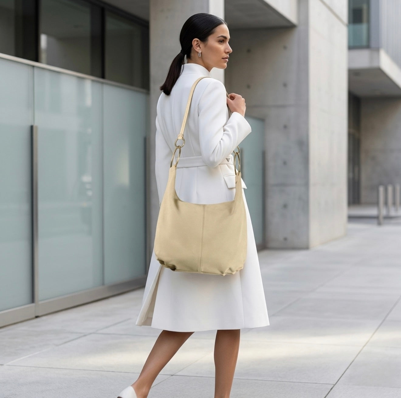 A woman in a white coat and heels walks outside a modern building, carrying a large handmade Italian bag over her shoulder.