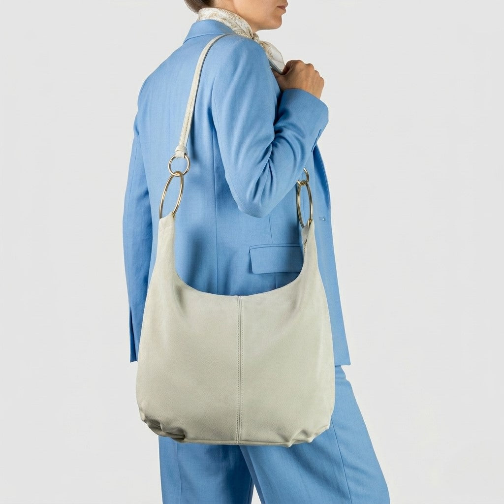 A person in a light blue suit and white scarf holds a large slouchy shoulder bag with metal rings, shown against a plain light background.