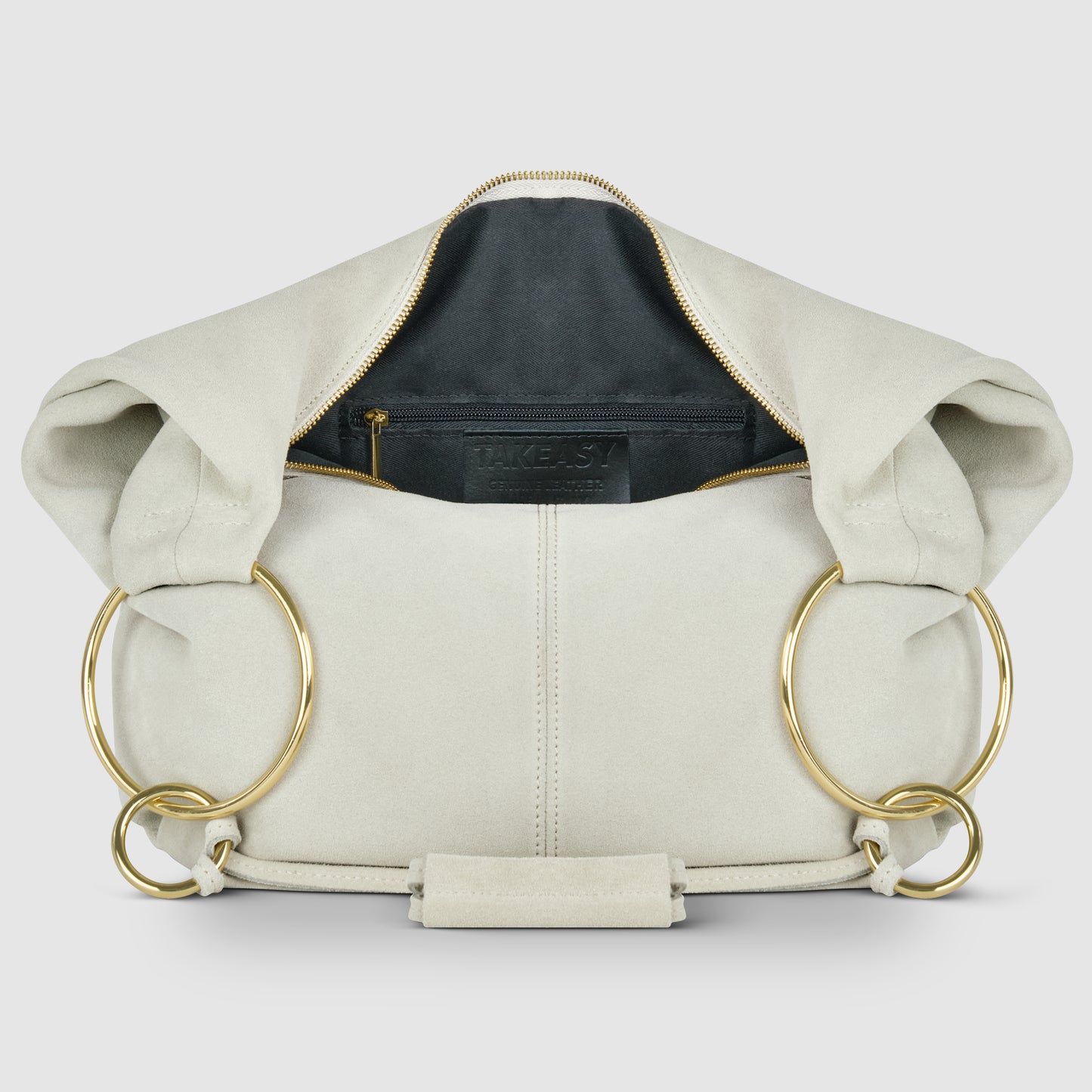 A cream-colored Italian handmade bag crafted from natural suede, with large gold rings as handles. Shown open to reveal a black interior lining and zippered pocket, the bag is displayed against a plain, light background.
