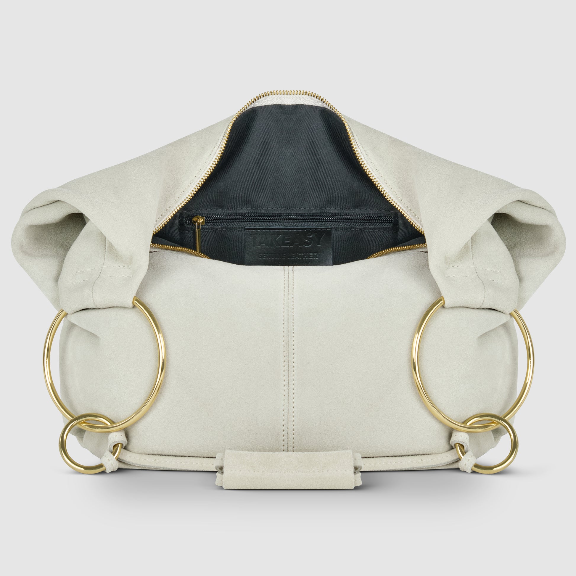 A cream-colored Italian handmade bag crafted from natural suede, with large gold rings as handles. Shown open to reveal a black interior lining and zippered pocket, the bag is displayed against a plain, light background.