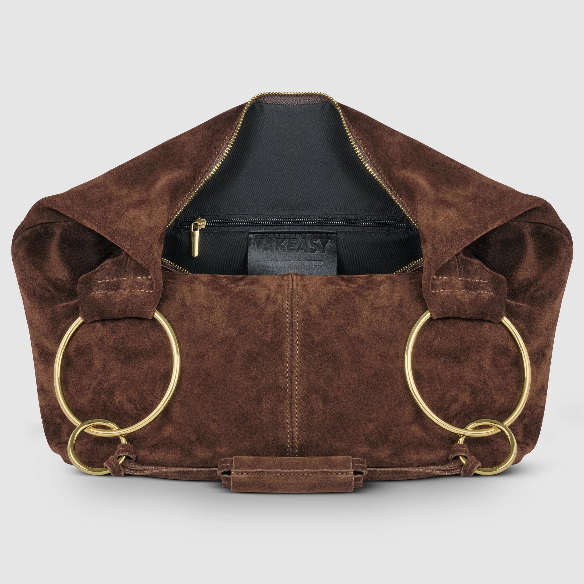 A natural suede bag with a zipper opening, large gold metal rings as handles, and a black inner pocket labeled TAKEASY. This Italian handmade bag is displayed against a plain light background.