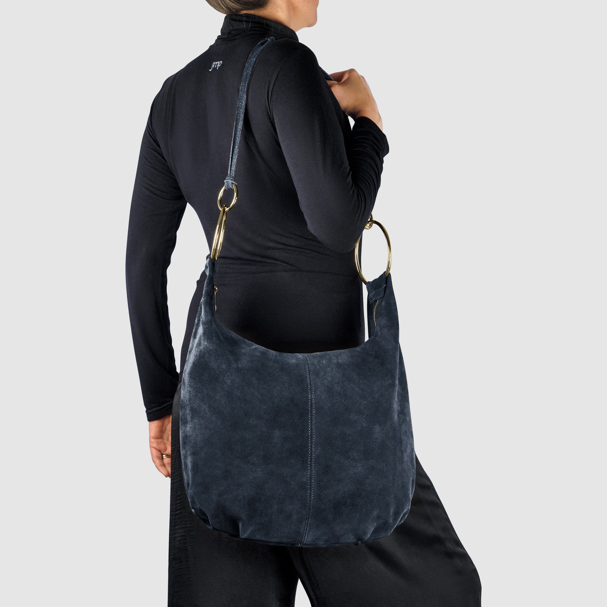 A person in black clothes is shown from the back, holding a large Italian handmade bag in blue natural suede with gold rings and a long strap, set against a plain grey background.