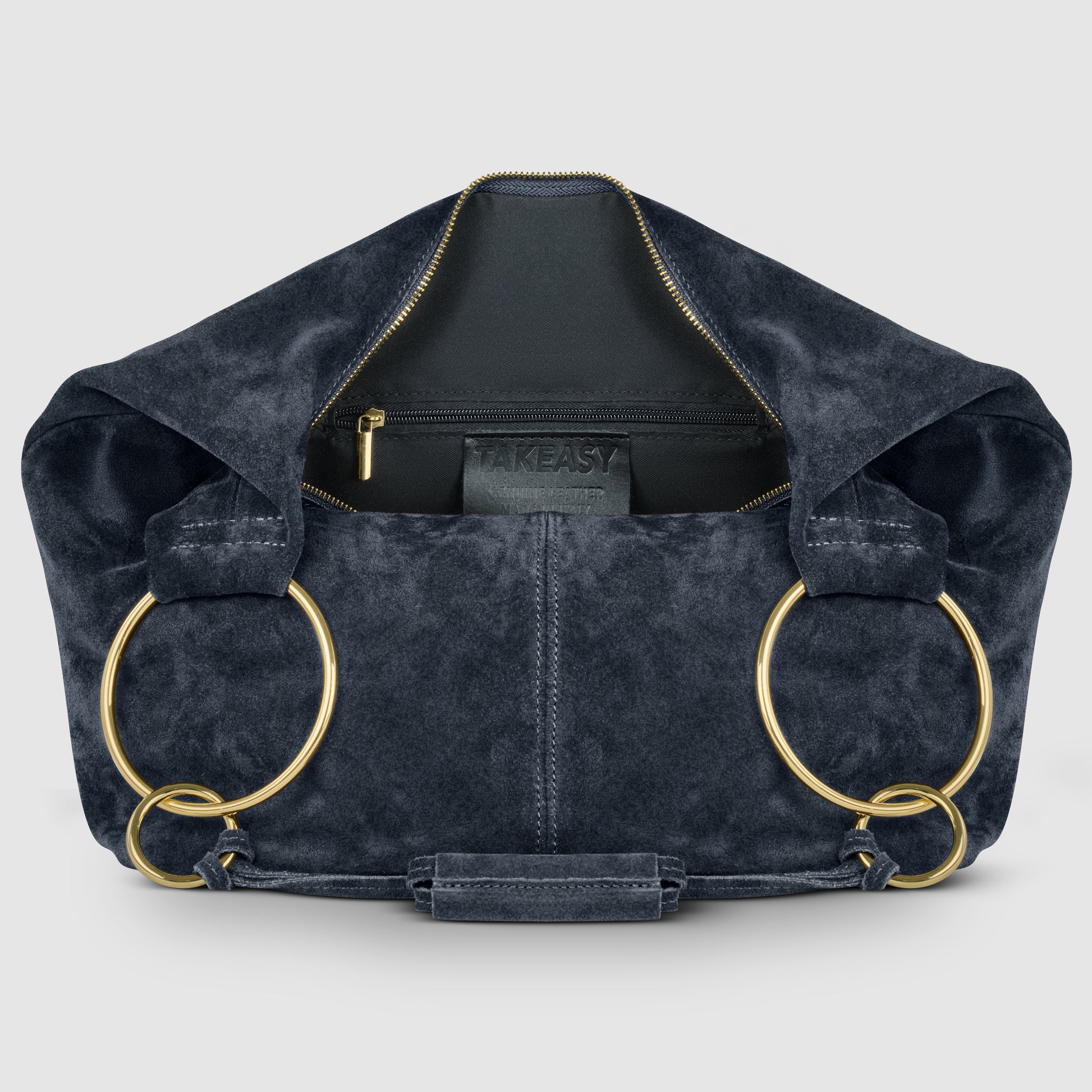 A dark blue Italian handmade bag crafted from natural suede, featuring large gold ring handles, an open zipper, and an inner pocket with a black TAKEASY label. The bag is displayed on a light gray background.