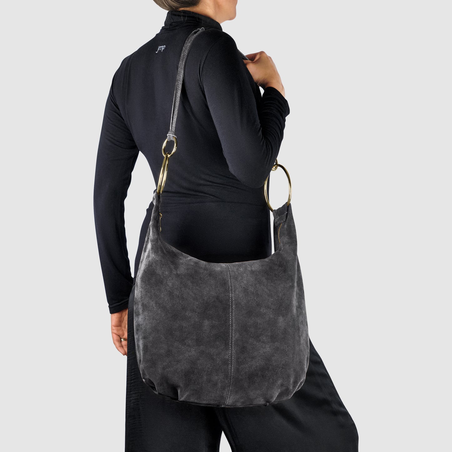 A person wearing a black long-sleeve top and pants holds an Italian handmade bag in dark gray natural suede with metal rings and an adjustable strap, shown against a plain light gray background.