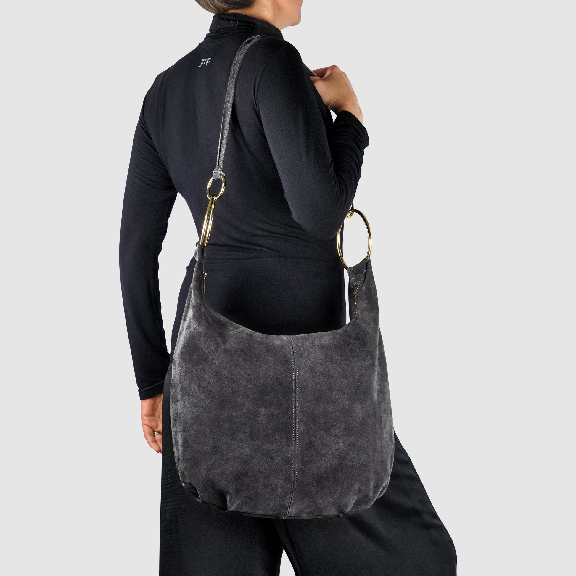 A person wearing a black long-sleeve top and pants holds an Italian handmade bag in dark gray natural suede with metal rings and an adjustable strap, shown against a plain light gray background.