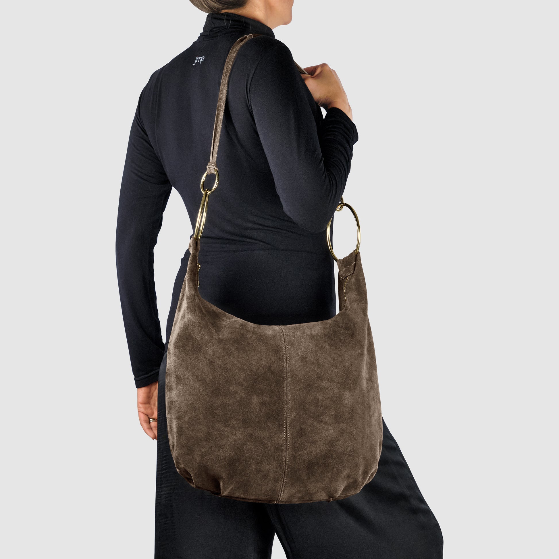 A person in a black outfit holds a large, Italian handmade suede leather bag with gold rings and a long strap, standing against a plain light gray background.