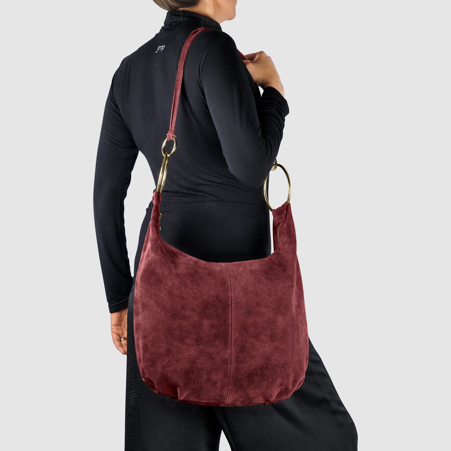 A person in a black long-sleeve top and pants carries a large, burgundy Italian handmade bag crafted from natural suede with gold metal rings and an adjustable strap against a light gray background.