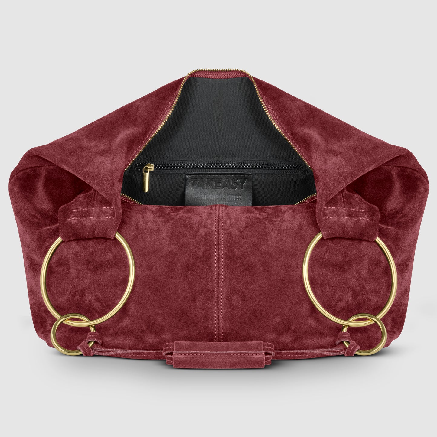 A burgundy Italian handmade bag in natural suede features large gold rings as handles, opening to reveal a black interior with a zippered pocket and visible TAKEASY label. The background is light gray.