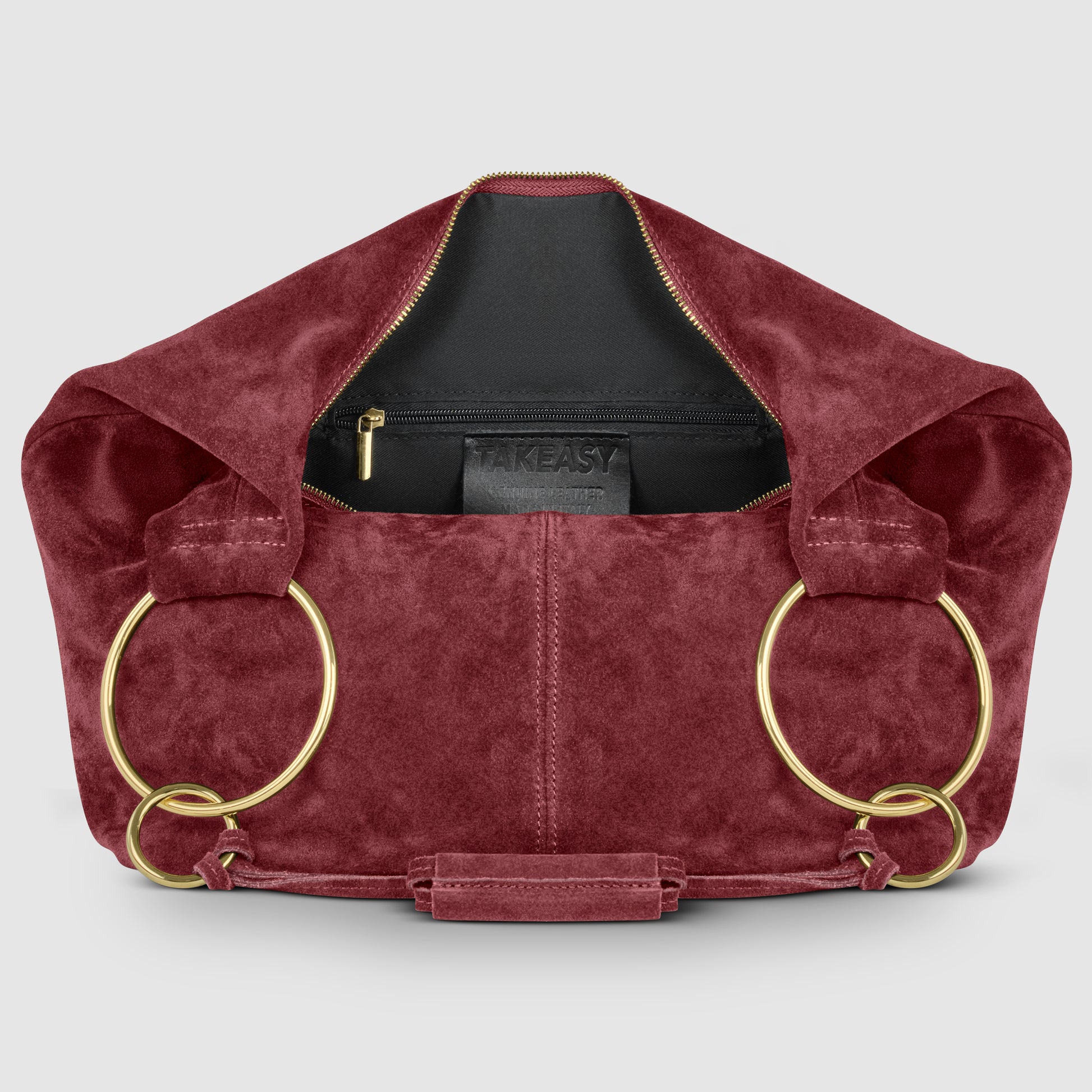 A burgundy Italian handmade bag in natural suede features large gold rings as handles, opening to reveal a black interior with a zippered pocket and visible TAKEASY label. The background is light gray.