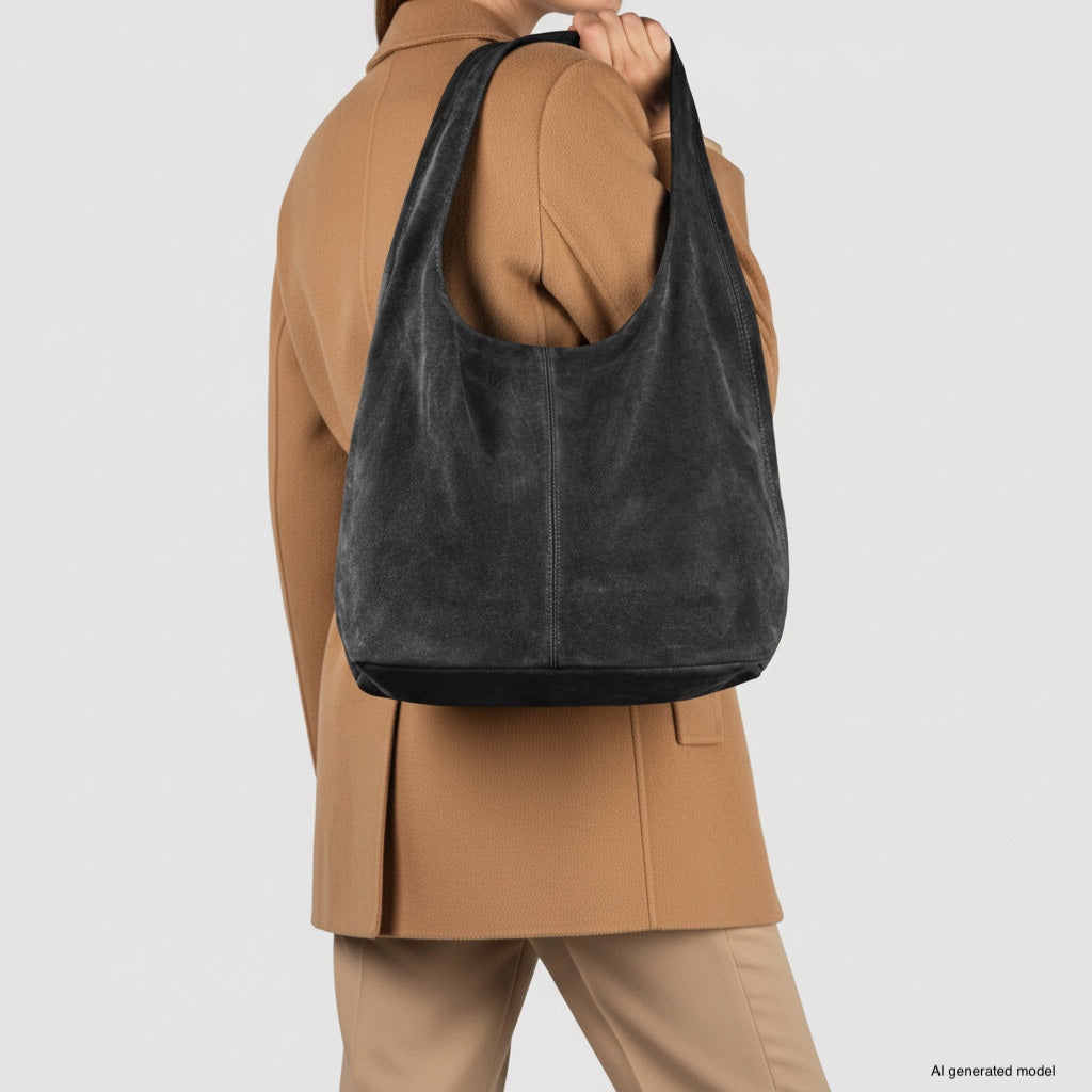 A person wearing a tan coat holds a slouchy suede shoulder purse. The spacious natural suede handbag is prominently displayed, with the individual’s head turned away from the camera against a plain background.