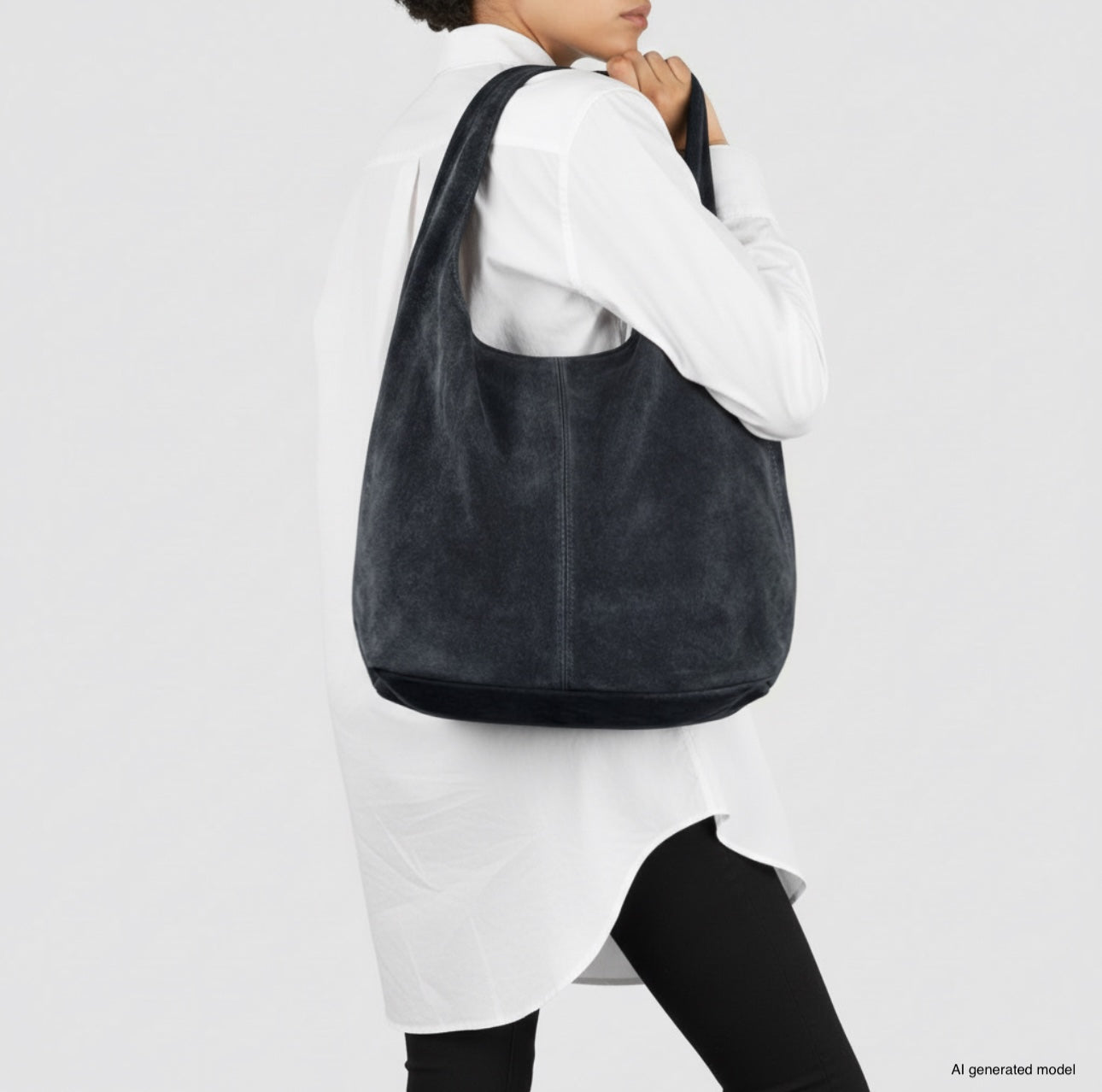 A person wearing a long white shirt and black pants carries a spacious natural suede handbag with a slouchy style. The background is plain and light-colored.