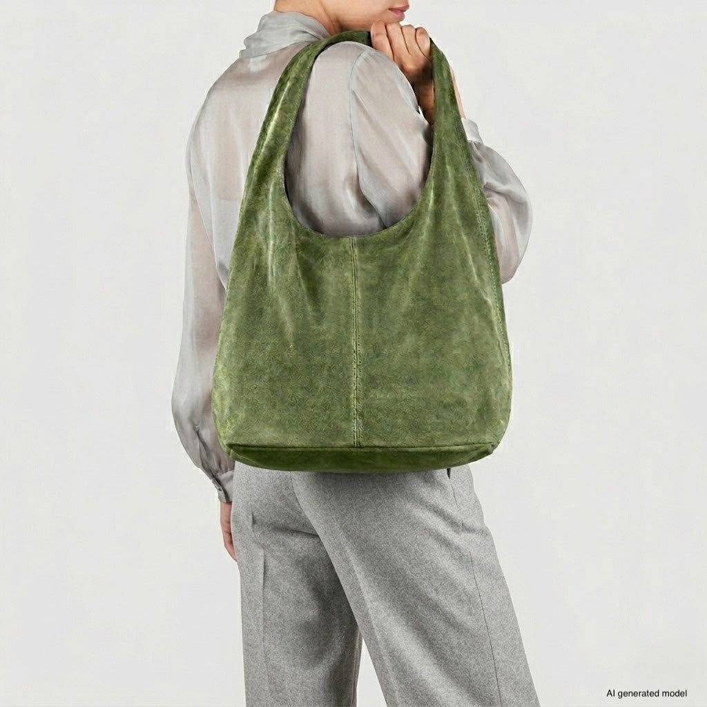 A person wearing gray pants and a sheer gray blouse holds a spacious natural suede handbag over their shoulder. The large, green hobo bag stands out against the plain, light-colored background.