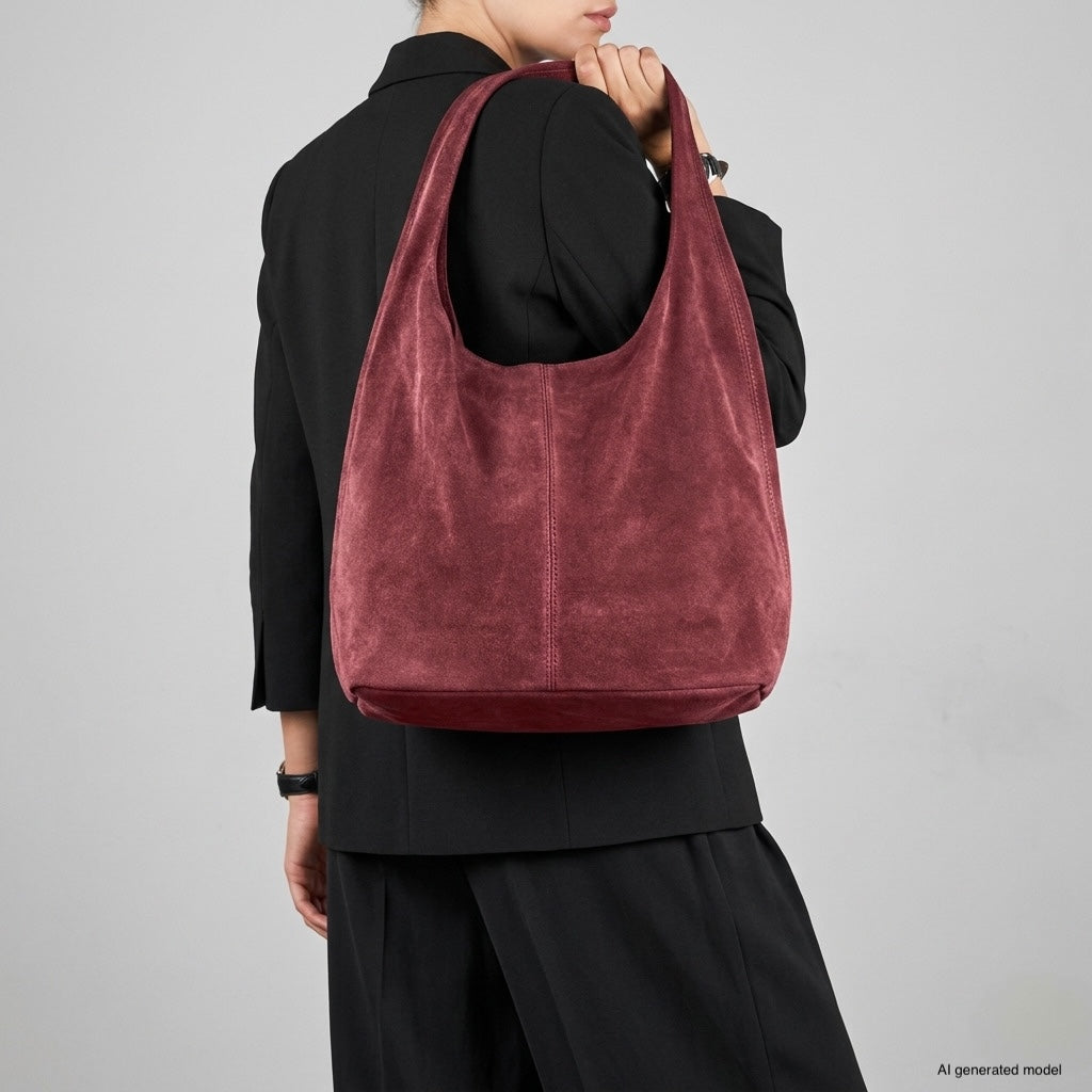 A person wearing a black blazer and trousers holds a slouchy suede shoulder purse in rich burgundy against a plain gray background. The spacious natural suede handbag hangs from their shoulder, with their face partially visible.