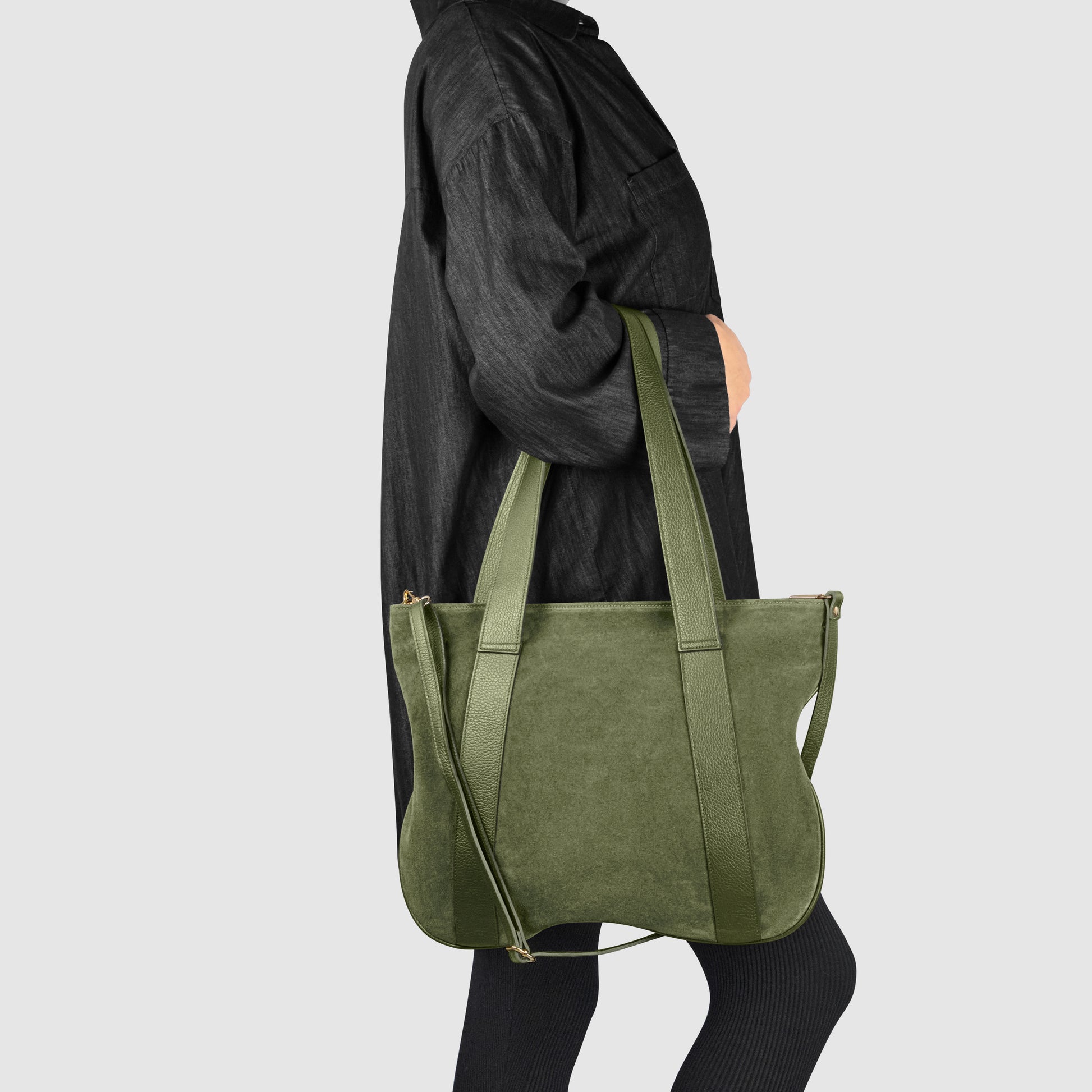 A person in a long-sleeve black outfit holds a handcrafted shoulder bag with two long straps, standing against a plain light gray background.