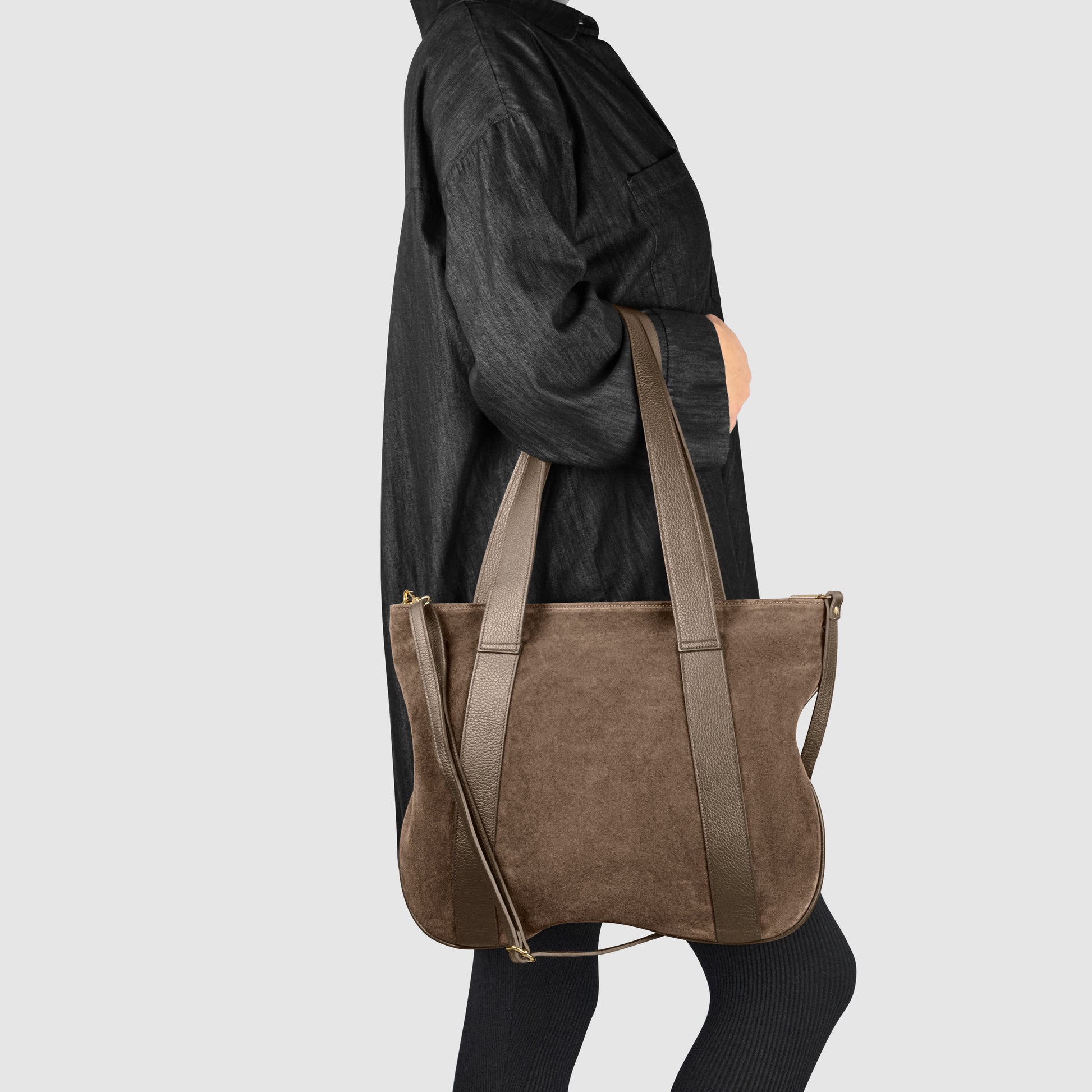 A person in a loose black outfit carries a large suede leather handbag with long leather straps over their shoulder, standing against a plain light gray background.