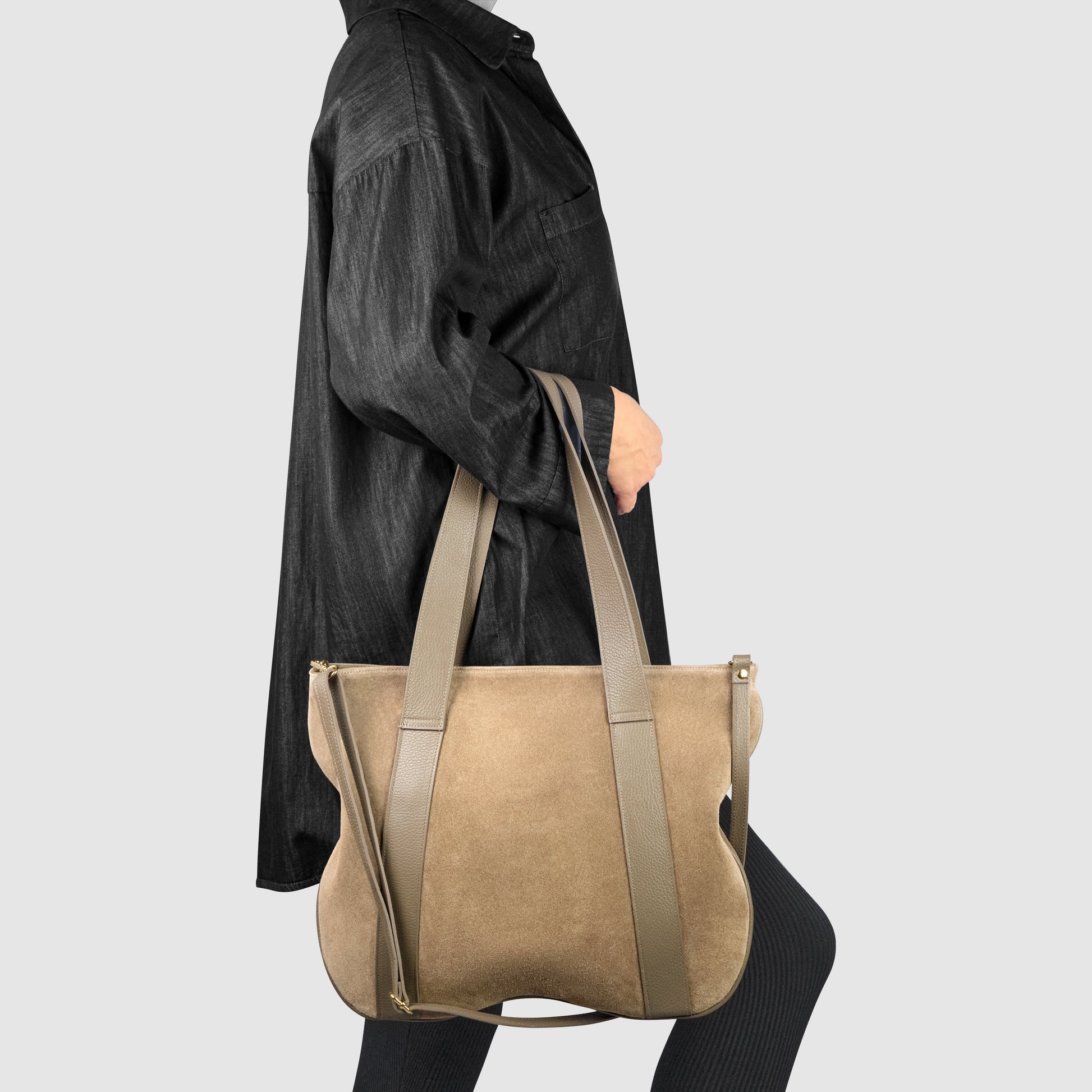 Person in a loose black shirt carrying a large beige suede leather handbag with wide straps over their shoulder, standing against a plain light grey background.