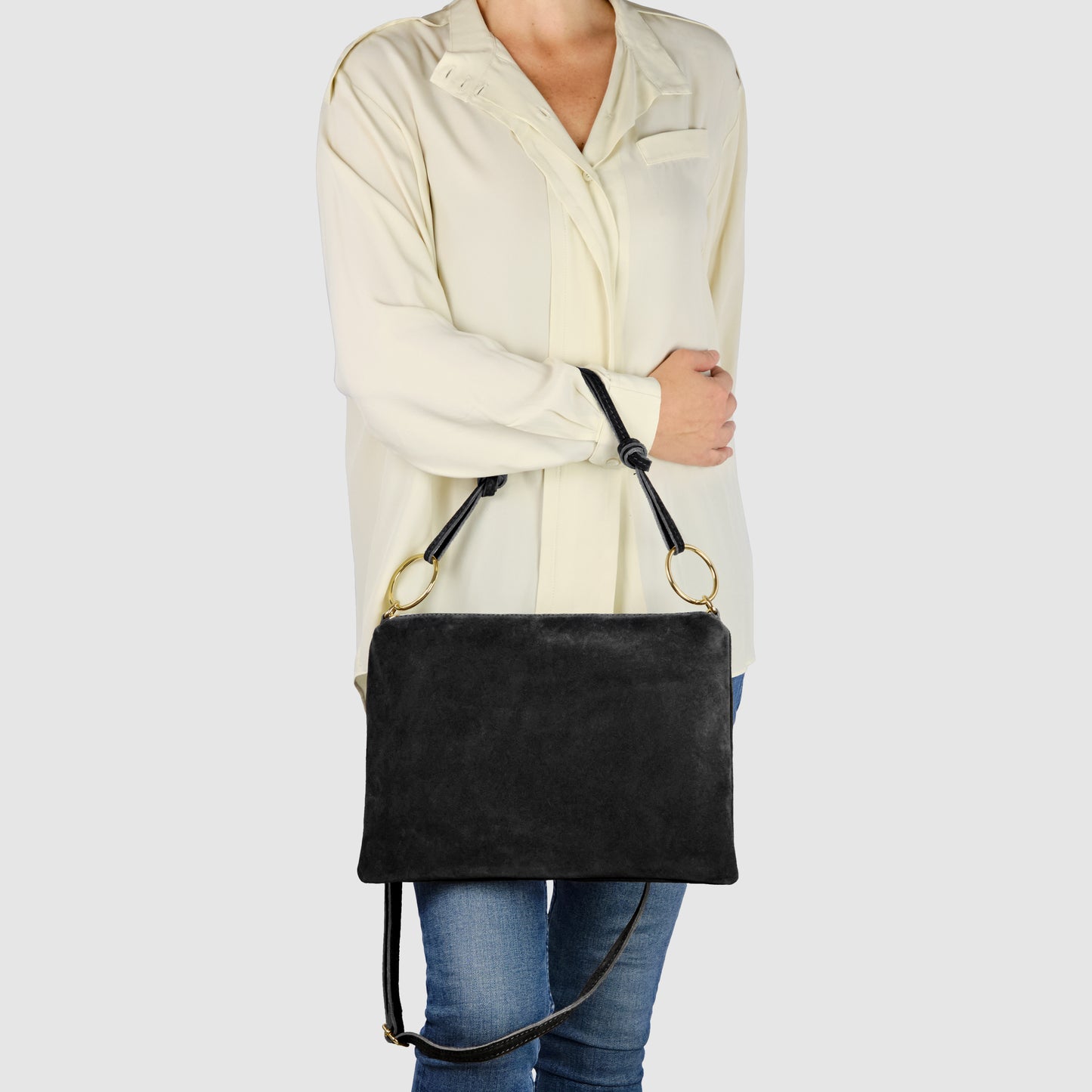 A person wearing a cream-colored blouse and blue jeans holds a real suede leather handbag with gold rings on the straps, standing against a plain light background.