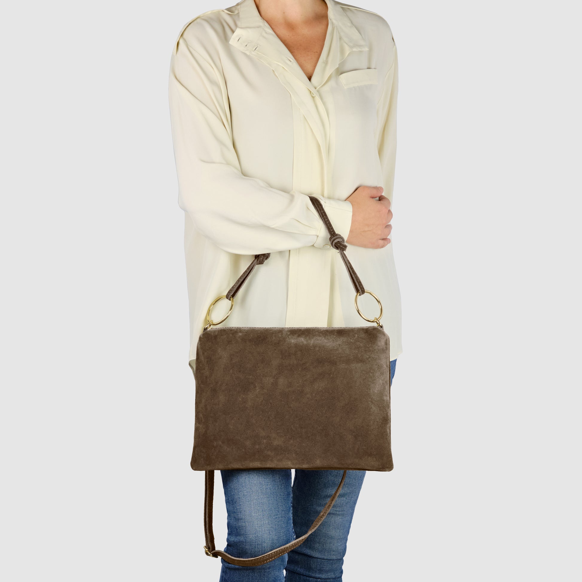 A person wearing a light beige blouse and blue jeans holds an Italian suede leather purse with gold rings, zip closure, and a shoulder strap against a plain light background.