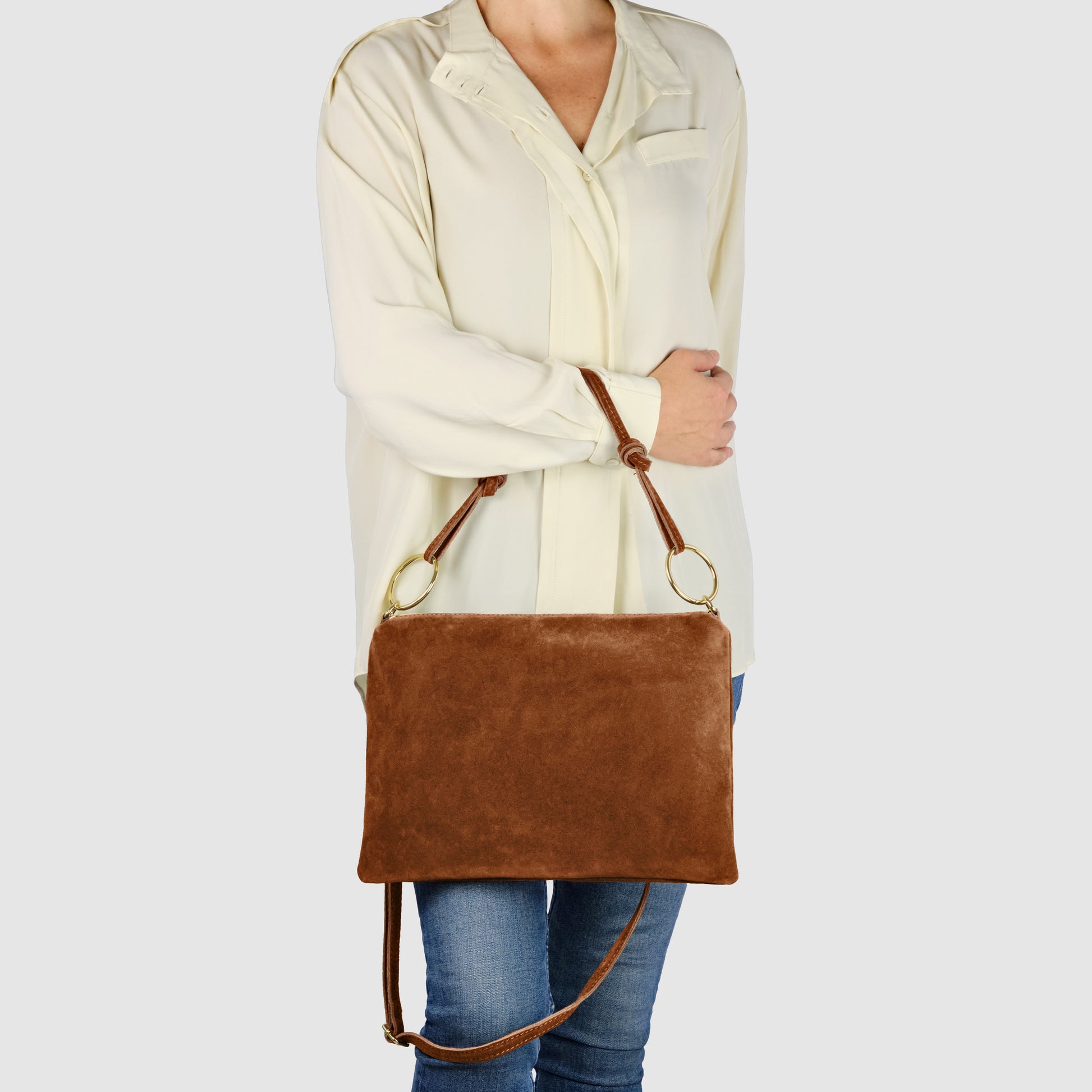 A person wearing a cream-colored blouse and blue jeans holds an Italian suede purse with gold rings and an adjustable strap against a plain light background. The medium zip closure purse is shown with the person’s head not visible.