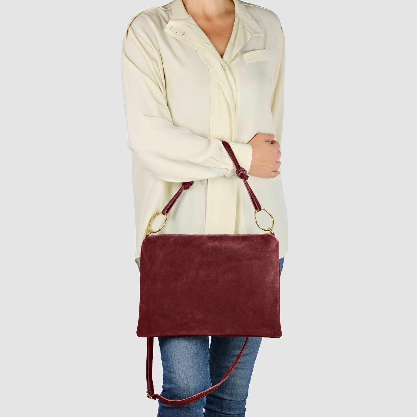 A person wearing a cream-colored blouse and blue jeans holds a slim medium purse—an Italian suede purse in maroon with gold rings and a shoulder strap—against a plain light background.