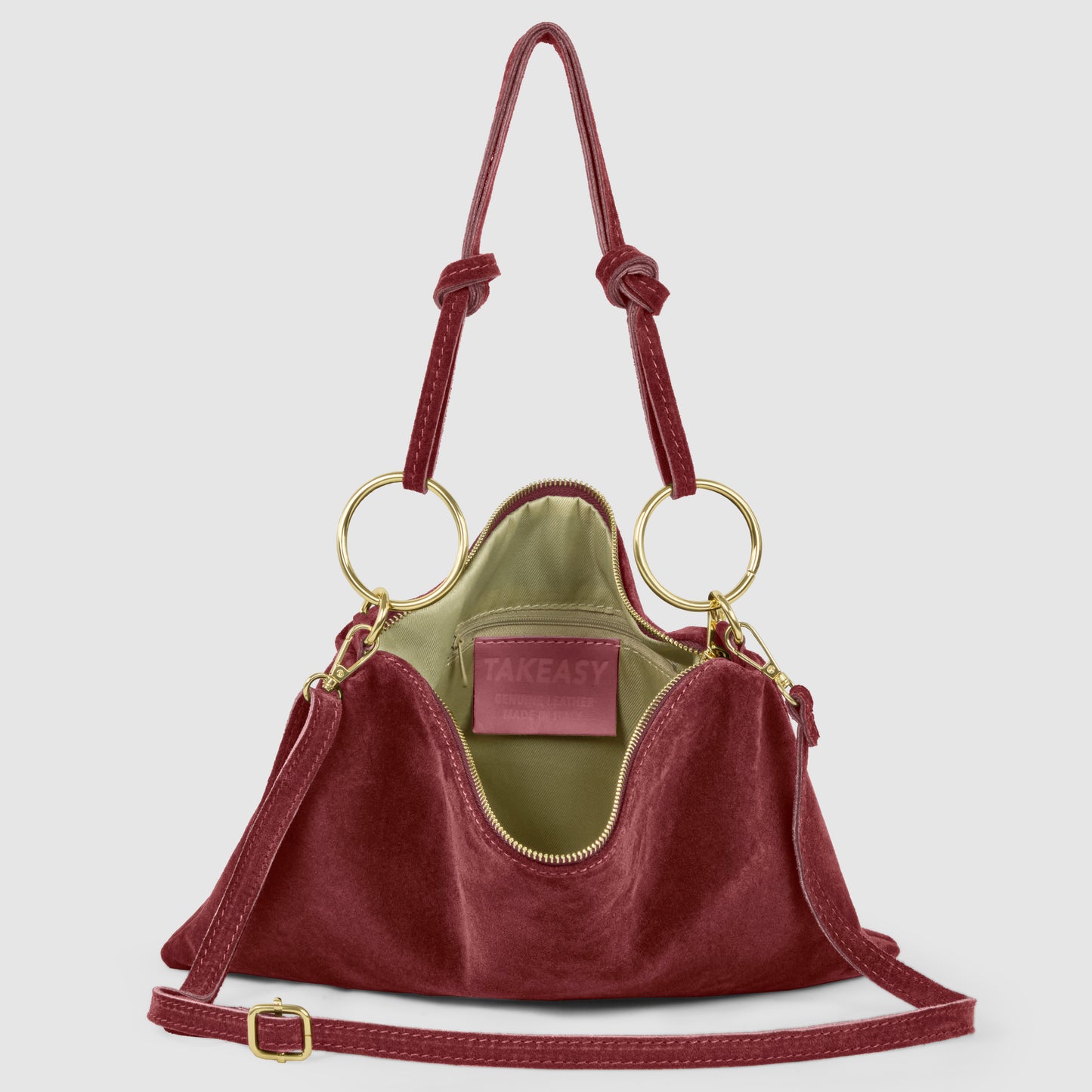 A slim medium purse in burgundy velvet with gold circular handles, a matching shoulder strap with gold hardware, and an olive green interior. Inside, a label reads TAKEASY.