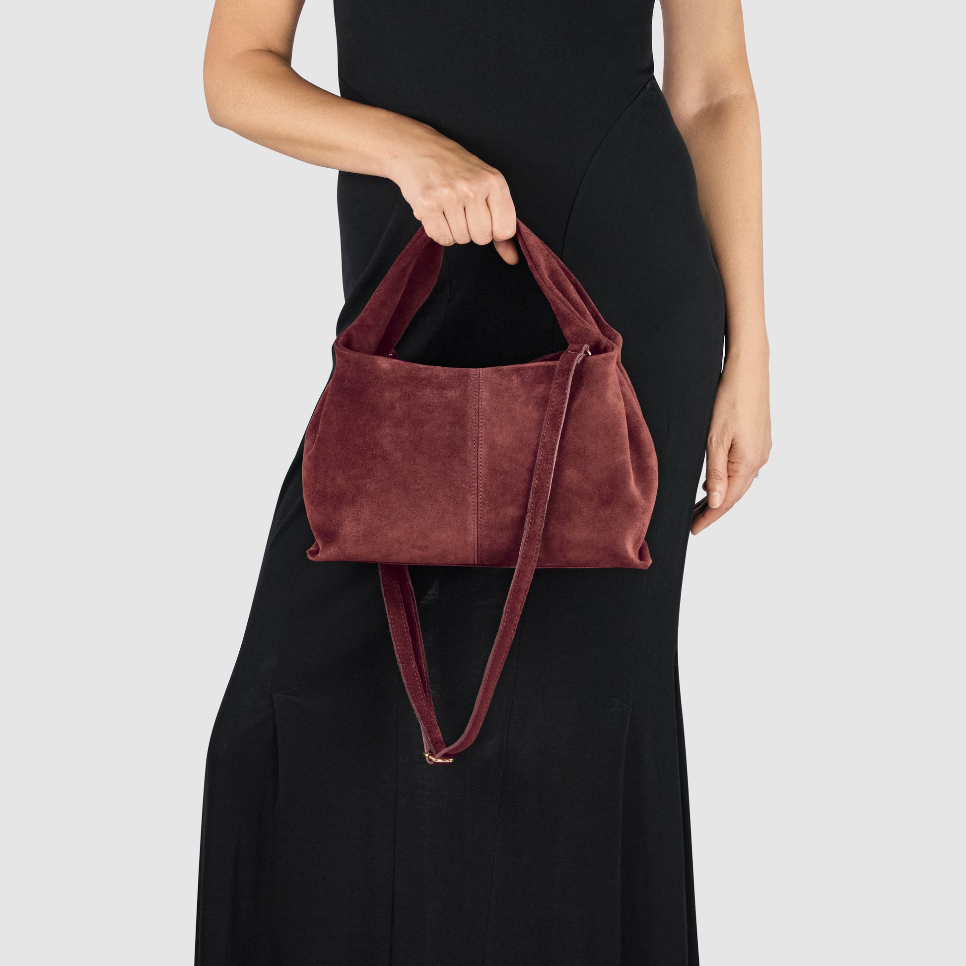 A woman in a sleeveless black dress holds a handcrafted suede bag with two handles and a shoulder strap, showcasing Florentine craftsmanship against a plain light gray background.