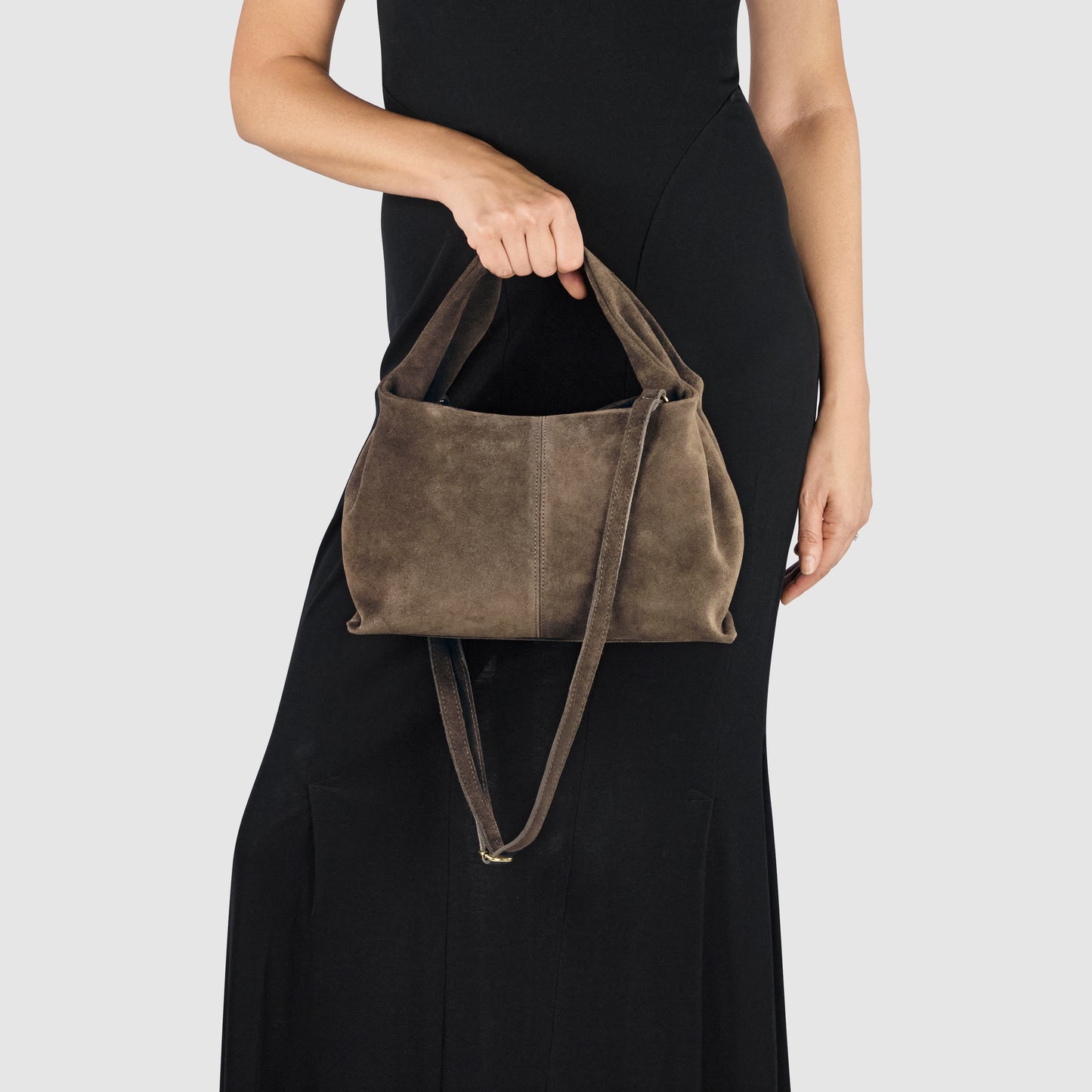 A person in a sleeveless black dress holds a handcrafted suede bag with two handles and a long strap, showcasing exceptional Florentine craftsmanship, against a plain light gray background.