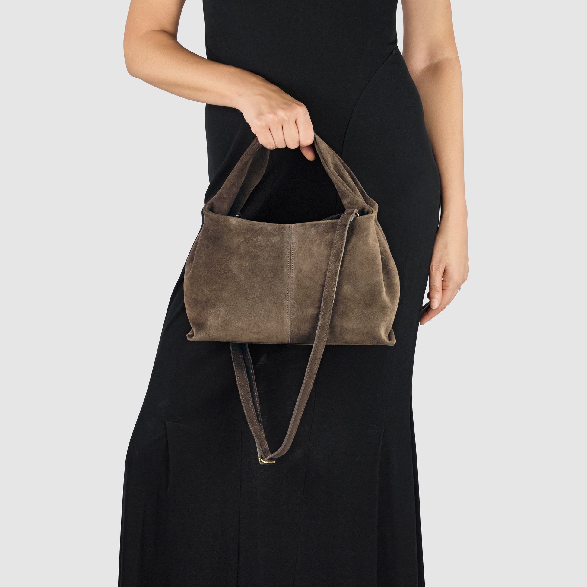 A person in a sleeveless black dress holds a handcrafted suede bag with two handles and a long strap, showcasing exceptional Florentine craftsmanship, against a plain light gray background.