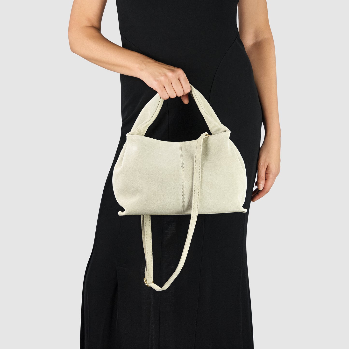 A person in a sleeveless black dress holds a handcrafted suede bag with both a short handle and a long shoulder strap, set against a plain light gray background.
