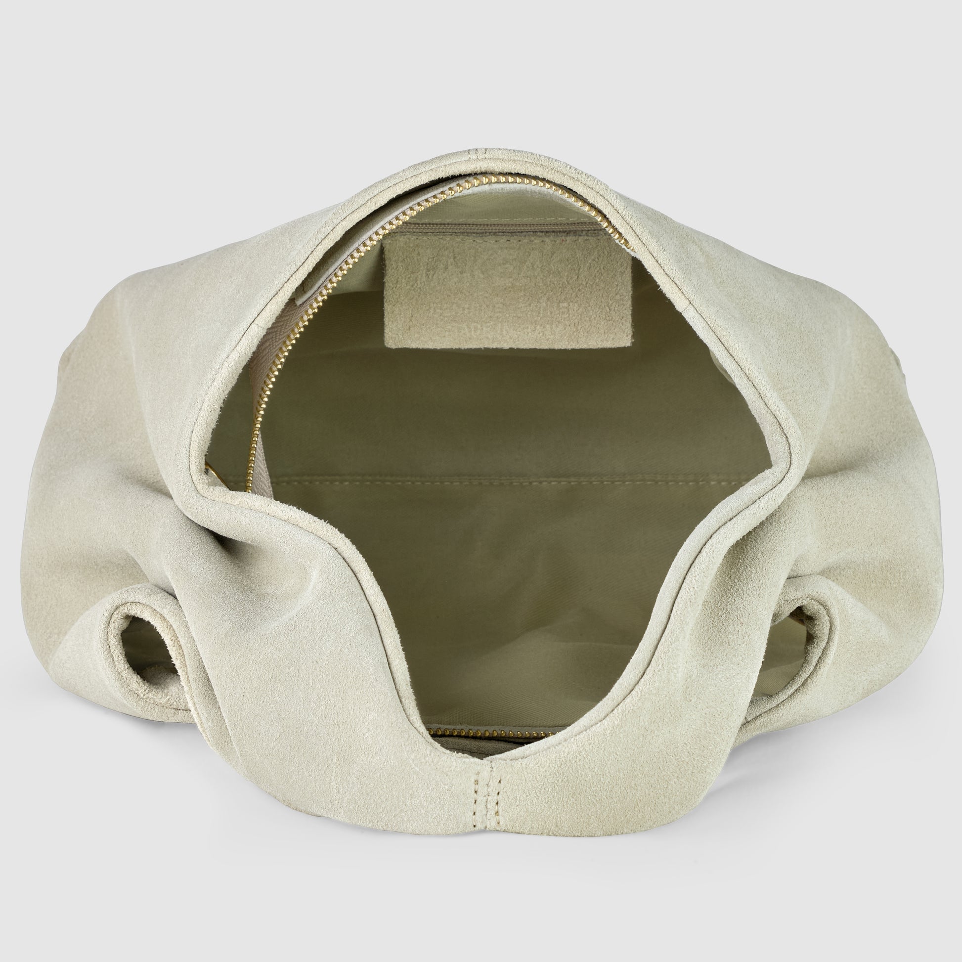 A handcrafted suede bag in beige is shown from above with its zipper open, revealing a spacious, empty interior and a small inner pocket on the back wall.