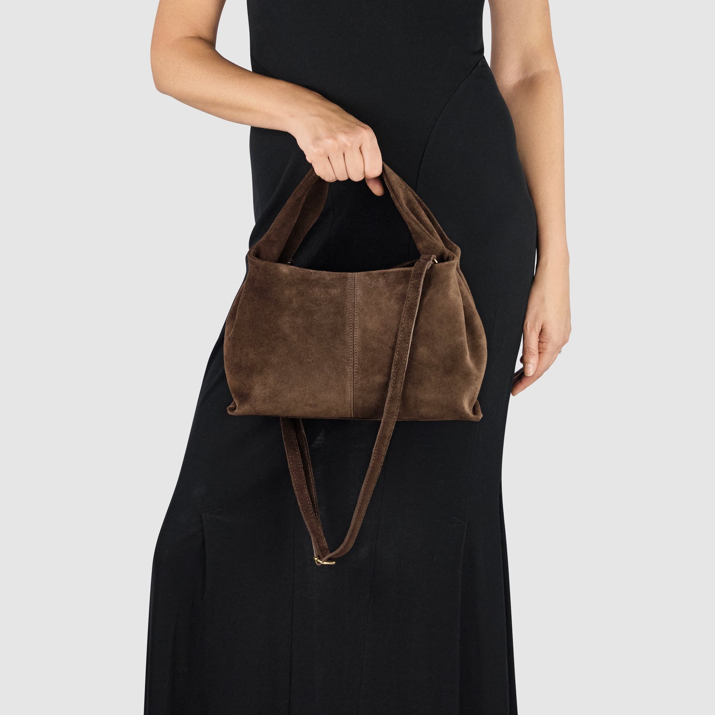 A person in a black sleeveless dress holds a large handcrafted suede bag with two handles and a long shoulder strap against a plain light background.